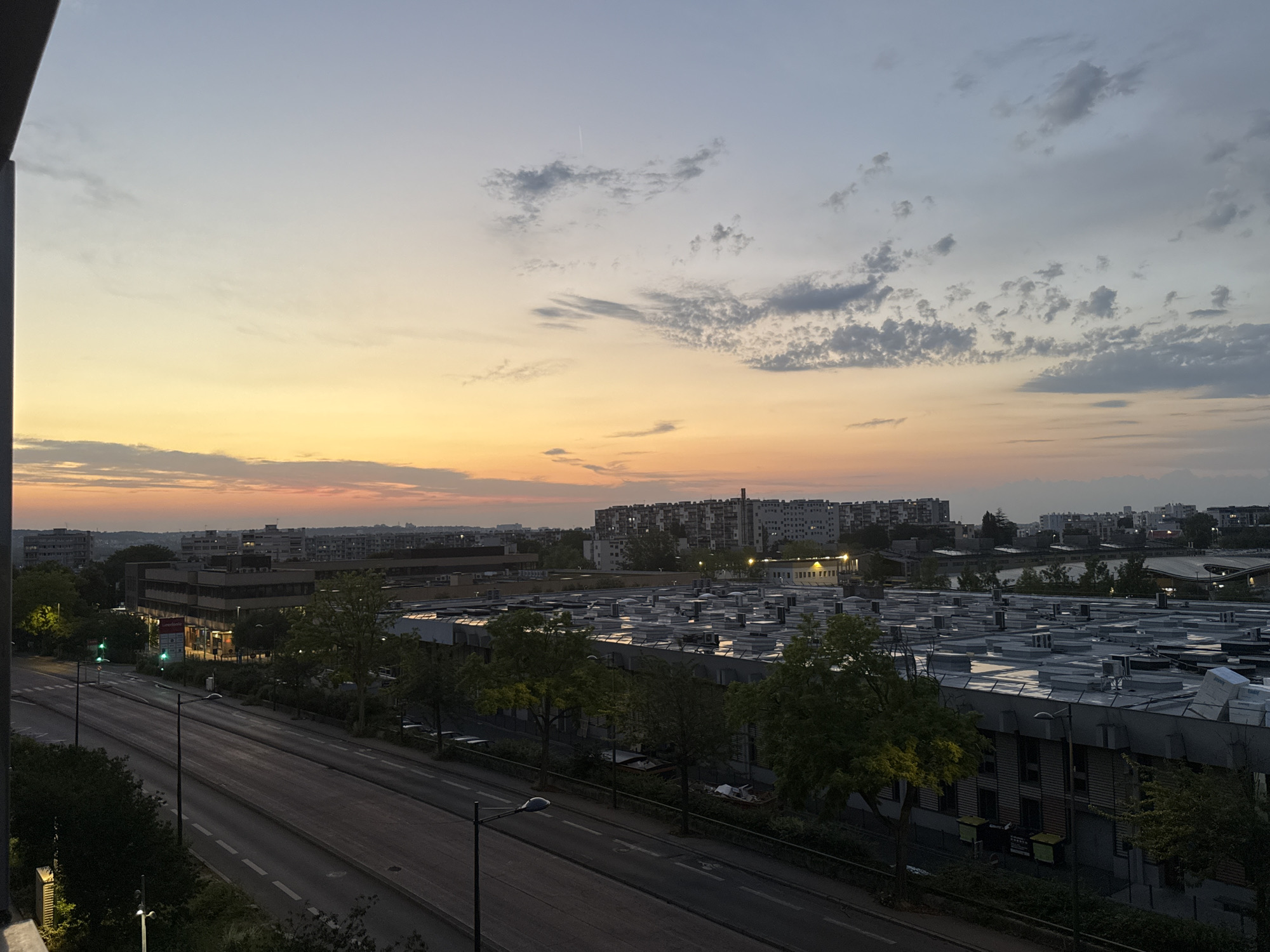 The image captures a cityscape at dusk, viewed from a slightly elevated perspective. The sky is a gradient of soft blues and oranges, with scattered clouds and a faint contrail visible in the upper left quadrant. Below the sky, a sprawling urban landscape unfolds, characterized by a mix of buildings of varying heights and architectural styles.
In the middle ground, a large, modern building with a flat, complex roof covered in numerous air conditioning units and vents dominates the scene. This building appears to be an industrial or commercial facility. Trees with green foliage line the front of this building, and some are illuminated by streetlights or the setting sun.
To the left of this large building, a street runs parallel to the foreground. It is a multi-lane road with clear lane markings and a few streetlights. Traffic lights are visible, showing green signals. On the far side of the street, there are smaller buildings, some with illuminated windows, suggesting commercial establishments.
In the background, a dense collection of residential buildings, characterized by apartment blocks, stretches across the horizon. Some of these buildings are lit, indicating evening activity. The overall impression is one of a typical urban environment at the end of the day. The lighting suggests the transition from daylight to evening, with artificial lights beginning to illuminate the scene.