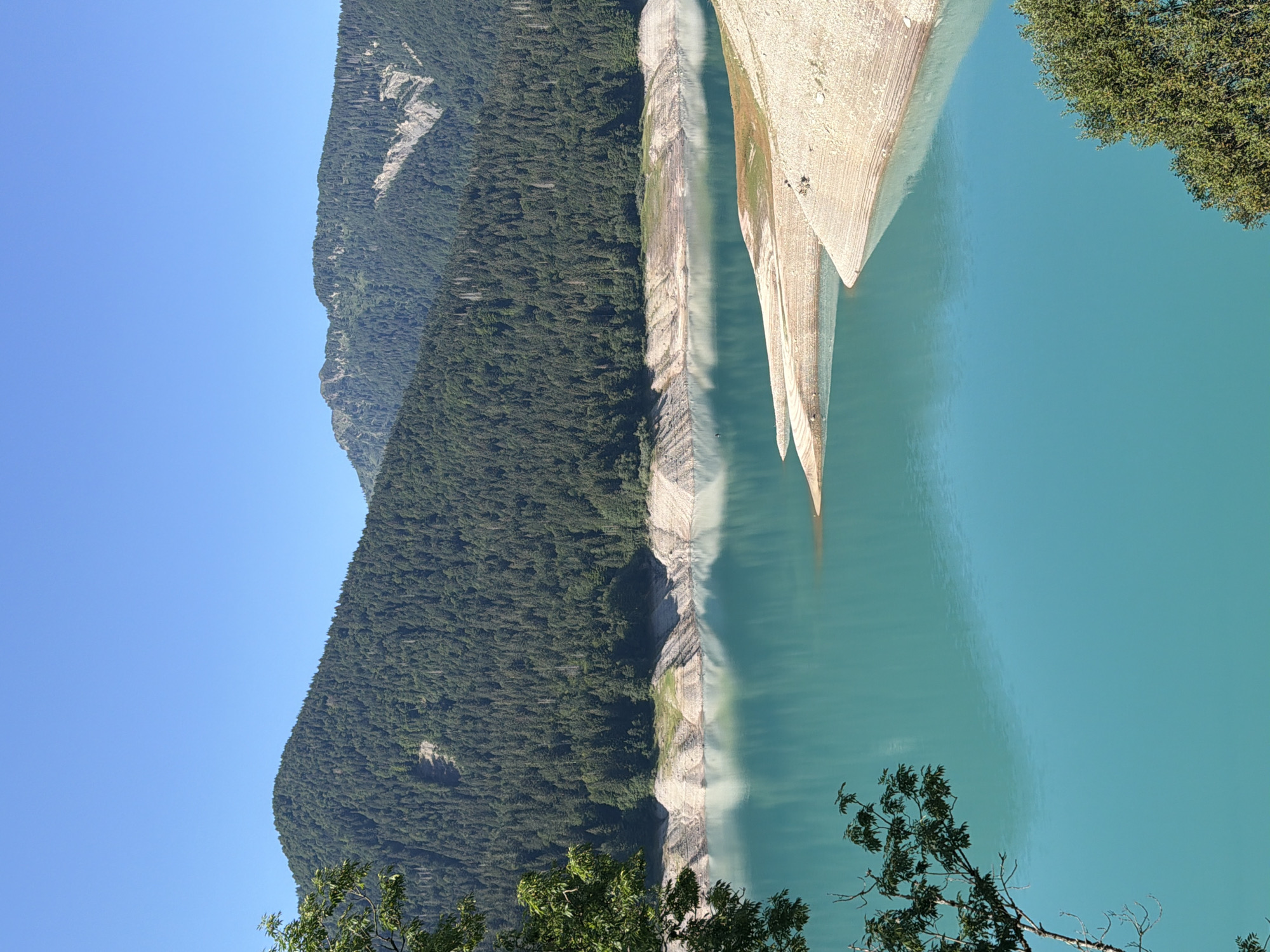A clear blue sky hangs over a tranquil, turquoise lake. The lake is surrounded by steep, forest-covered mountains that rise sharply from the water's edge. The lower slopes of the mountains are densely packed with evergreen trees, while the upper reaches are rocky and bare. The shoreline of the lake is rocky and uneven, with visible layers of sediment and erosion. A small, sandy peninsula juts out into the water on the right side of the image. The water reflects the blue of the sky and the green of the trees. The image captures a serene natural landscape, likely a reservoir or a mountain lake.