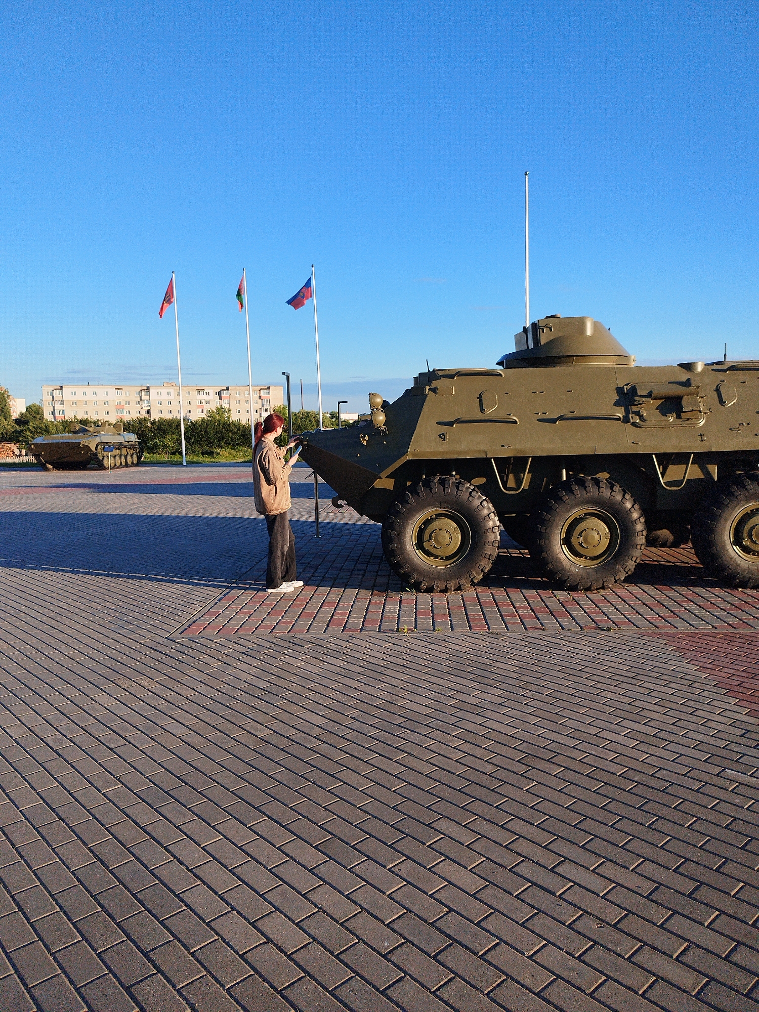 A young woman with red hair, wearing a brown jacket and dark pants, stands in front of a parked military vehicle. The woman is facing away from the camera, looking at a screen in her hand. The military vehicle is large and green, with multiple wheels. In the background, there are flags and buildings. The scene appears to be outdoors on a paved area.