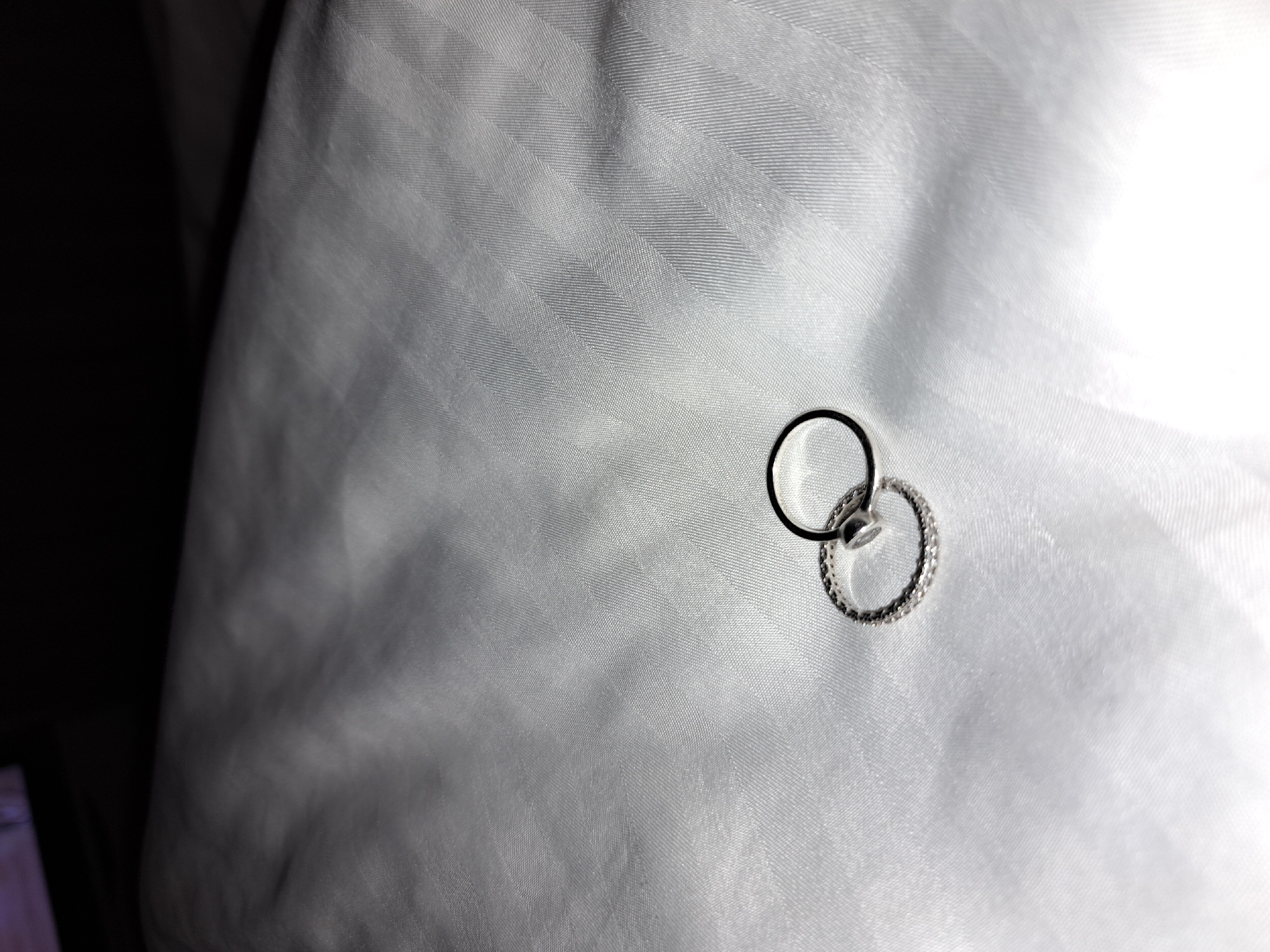 The image shows two rings resting on a white, textured fabric surface. One ring is a simple, plain band, while the other is decorated with small, possibly encrusted, stones. The background consists of the same white fabric, with a darker area visible in the upper left corner. There are no people or identifiable locations within the image. There is no indication that this image was taken in the city of Negovan, Bulgaria.