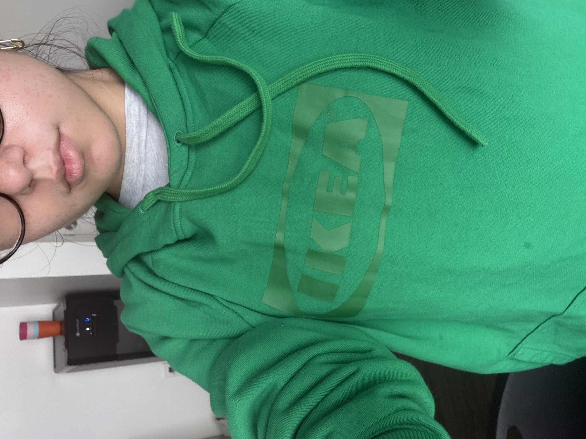 A person is wearing a green hoodie with the IKEA logo. The background shows a wall, and what appears to be a small electronic device with a pink and orange object attached to it. It is not possible to determine the exact location in Austin, United States of America from the provided image.