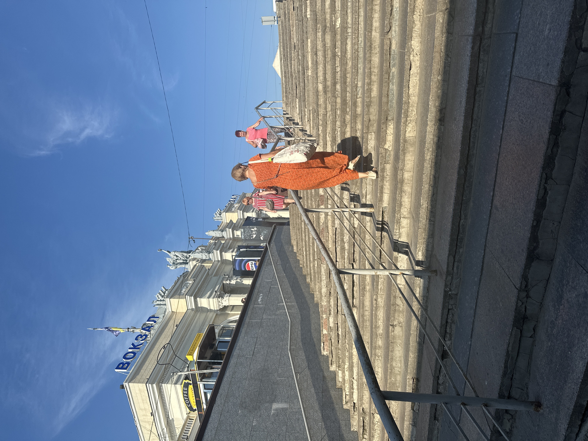 The image depicts the outdoor staircase leading up to the Odesa Railway Station, commonly referred to as "Vokzal" in Ukrainian, which is prominently displayed on the building's facade. The sky is clear blue with a few wispy clouds. Three women are visible ascending the wide, worn stone steps. The woman in the foreground wears an orange patterned dress and carries a large, light-colored tote bag. To her left, further up the stairs, a woman in a striped dress and a woman in a pink tank top are also climbing. A metal handrail runs along the side of the stairs. The architecture of the railway station is visible in the background, featuring classical columns and decorative sculptures. A coffee stand is also visible on a platform above the stairs. The overall scene suggests a sunny day at a bustling transportation hub in Odesa, Ukraine.