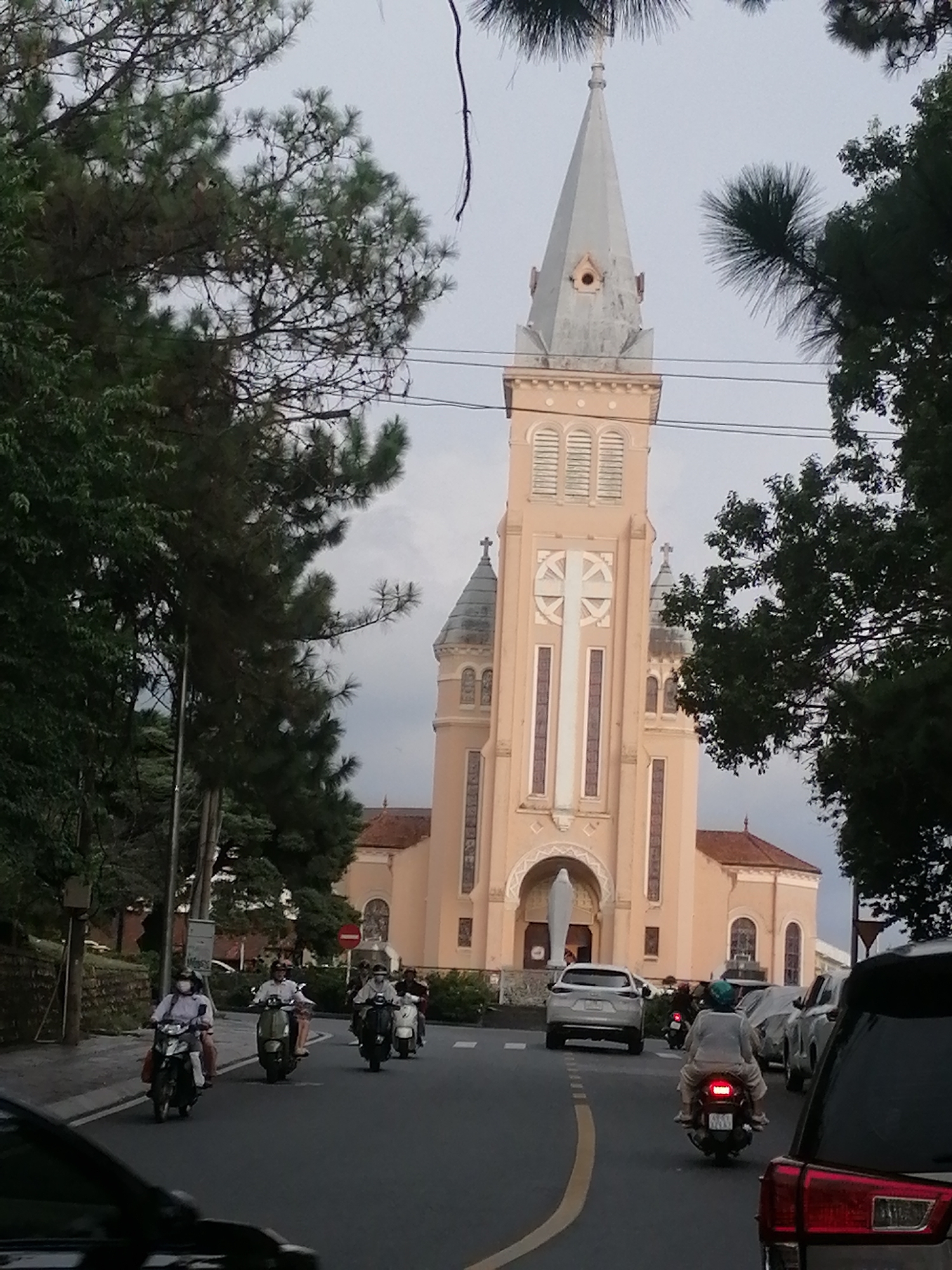 This is a slightly elevated, eye-level, medium shot of a street scene in Da Lat, Vietnam, featuring a prominent church with a tall spire. The church is a pale pink color and has architectural details including arched windows, crosses, and a large cross emblem on its facade. Trees with dark green foliage frame the church, partially obscuring the sky, which is overcast. In the foreground, a road is visible with several motorcycles and cars. Three people on motorcycles are in the middle ground, two wearing masks. One motorcycle has a person in a white shirt and another in a dark shirt. Another motorcycle to the right has two people on it, one wearing a blue helmet. In the distance, several cars and motorcycles are also visible. The road is paved and marked with white lines.
