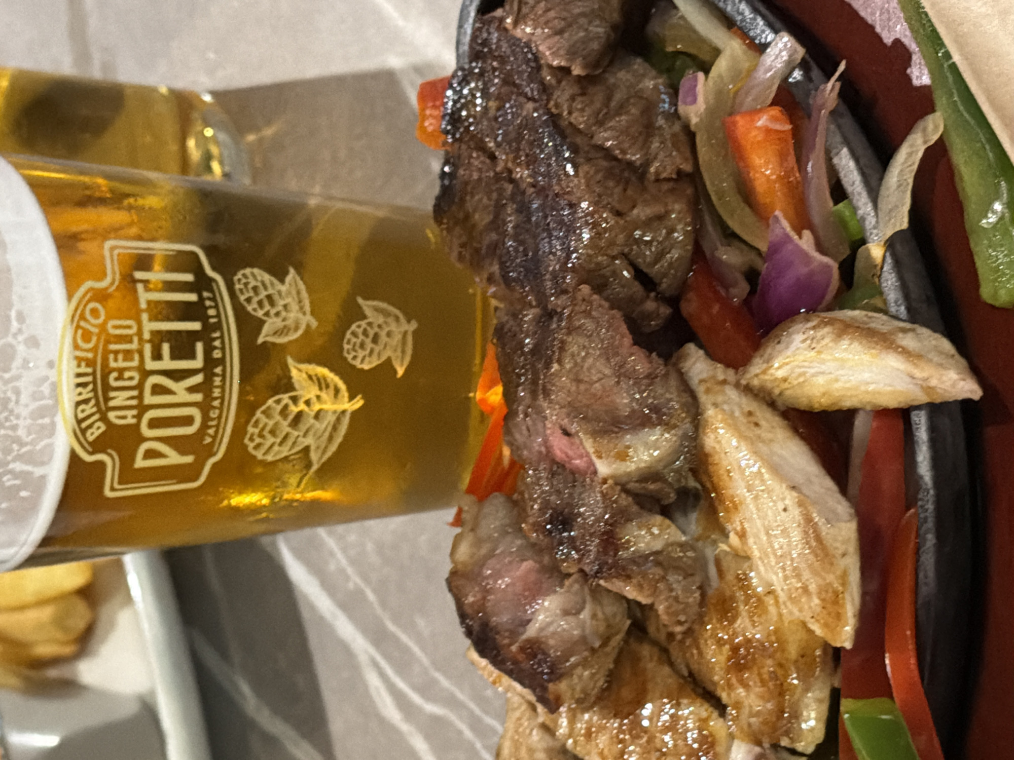 This close-up image, captured in Milan, Italy, features a meal being served on a tabletop. The primary subjects are a glass of beer and a sizzling platter of food.
The beer glass, occupying the left half of the frame, is filled with a clear, golden-amber liquid, indicating a lager or pale ale. The glass prominently displays a white logo with the text "BIRRIFICIO ANGELO PORETTI" at the top, and "VALGANNA DAL 1877" below, along with illustrations of hop cones. The glass appears frosted or has condensation, suggesting the beer is cold.
To the right, a dark, presumably cast-iron, platter holds a mix of cooked ingredients. Sliced pieces of beef, showing some charring and glistening surface moisture, are interspersed with strips of what appears to be grilled chicken. The meat and chicken are accompanied by colorful sautéed vegetables, including red and green bell peppers, orange bell peppers, and purple-red onions. The food looks freshly cooked and hot, with a glossy sheen from juices or oil.
In the background, a hint of fried potato items, possibly French fries, can be seen, suggesting a side dish to the main meal. The table surface is light-colored, possibly white or light grey, with a subtle textured pattern. The lighting is warm and artificial, typical of an indoor restaurant or bar setting, likely indicating an evening meal. The scene captures the moment of enjoying a hearty meal with a cold beer.