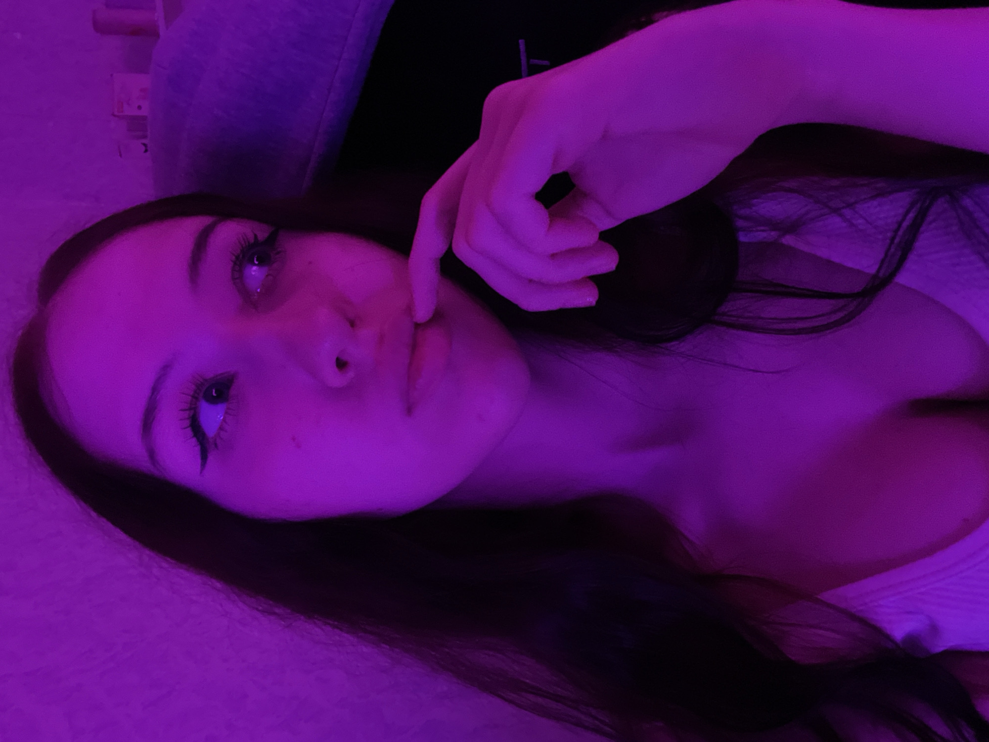 The image shows a young woman illuminated by purple light. She has long dark hair and is wearing a white top. Her left hand is raised to her mouth, with her index finger touching her lips. Her eyes are looking to her right. The background is a textured wall and a purple chair or cushion. There are no identifiable landmarks or features that would allow for an accurate location within the city of Votkinsk, Russia.