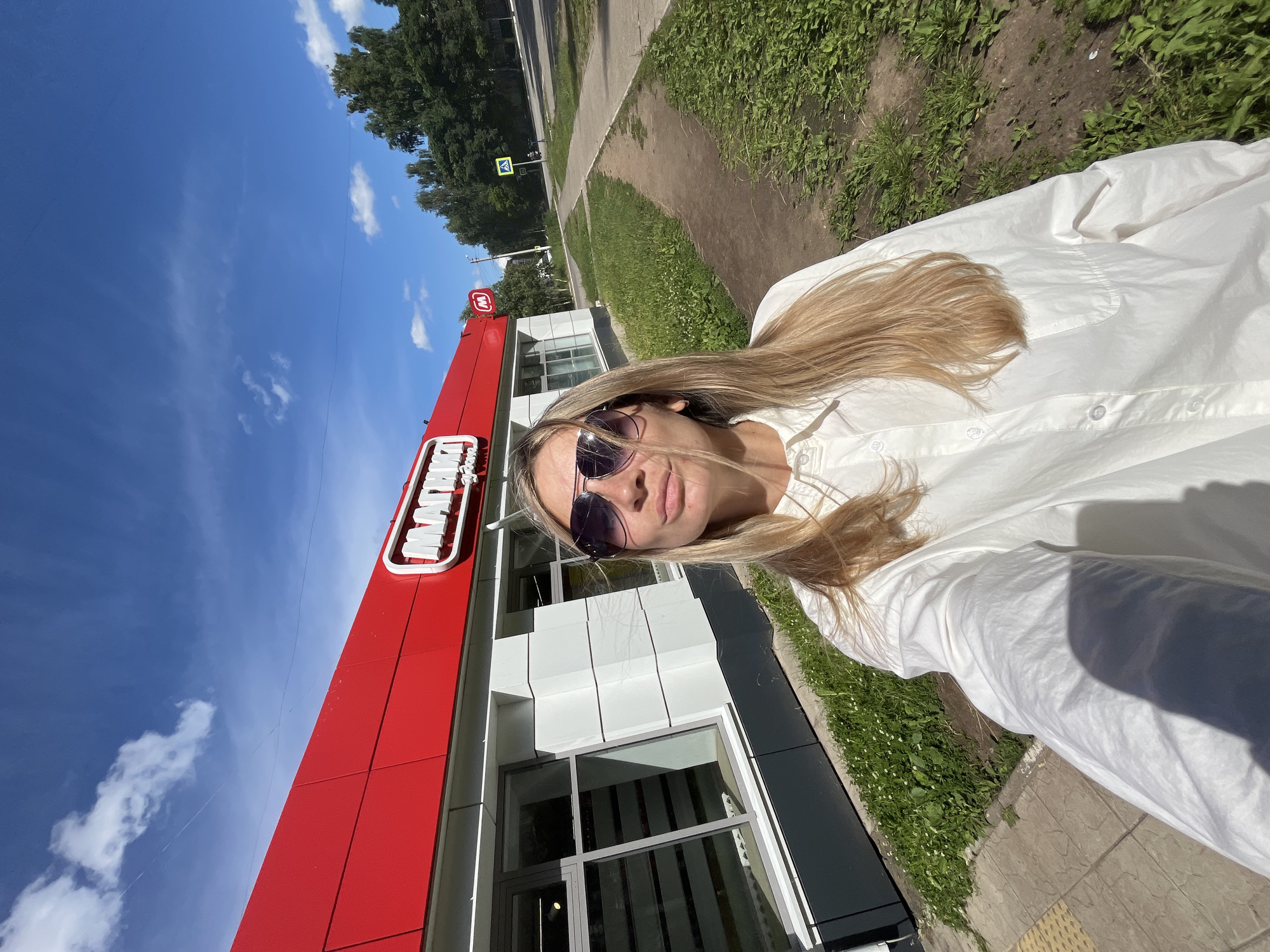A young woman with long blonde hair and sunglasses is taking a selfie outdoors. She is wearing a white button-down shirt. The woman is standing in front of a building with a bright red facade and white trim. A sign with white lettering on a red background is visible on the building. The background shows a sidewalk, green grass, trees, and a blue sky with some white clouds. The sun is shining brightly, indicating it is daytime. The image appears to be taken on a sunny day. The overall mood is casual and relaxed. The visible text on the sign appears to be "МАГНИТ".