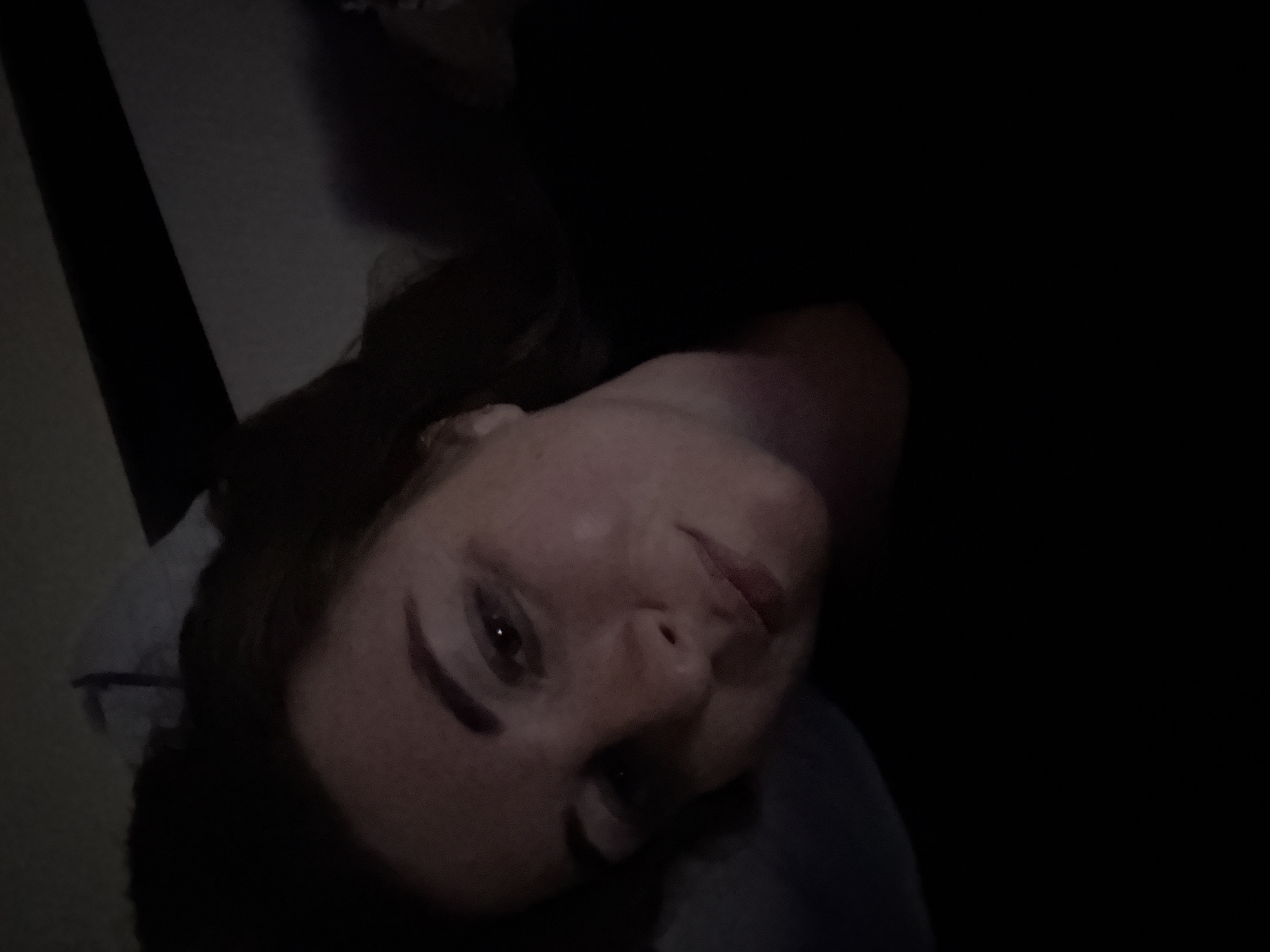A young woman is captured in a close-up, low-light selfie. Her head is tilted, and her gaze is directed towards the camera with a slight smile. She has fair skin and reddish-brown hair that frames her face. She is wearing a dark-colored top. The background is dark and indistinct, suggesting an indoor setting, likely a bedroom. A pillow is visible behind her head, partially obscured by her hair. The overall lighting is dim, with the light source seemingly coming from above and to the side, highlighting her facial features.