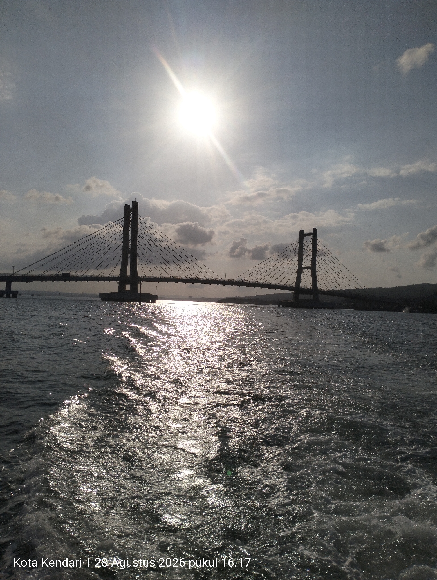 The image shows a wide shot of a cable-stayed bridge spanning across a body of water under a bright, sunlit sky. The sun is positioned high in the sky, creating a strong lens flare and reflecting brightly off the water's surface, forming a shimmering path. The bridge has two distinctive towers, each supporting a network of cables that hold up the road deck. The water in the foreground exhibits a wake, suggesting movement, possibly from a boat. The sky is partially filled with scattered clouds. A timestamp and location are visible in the bottom left corner: "Kota Kendari | 28 Agustus 2026 pukul 16.17". The scene depicts a large infrastructure project within an urban or coastal environment.