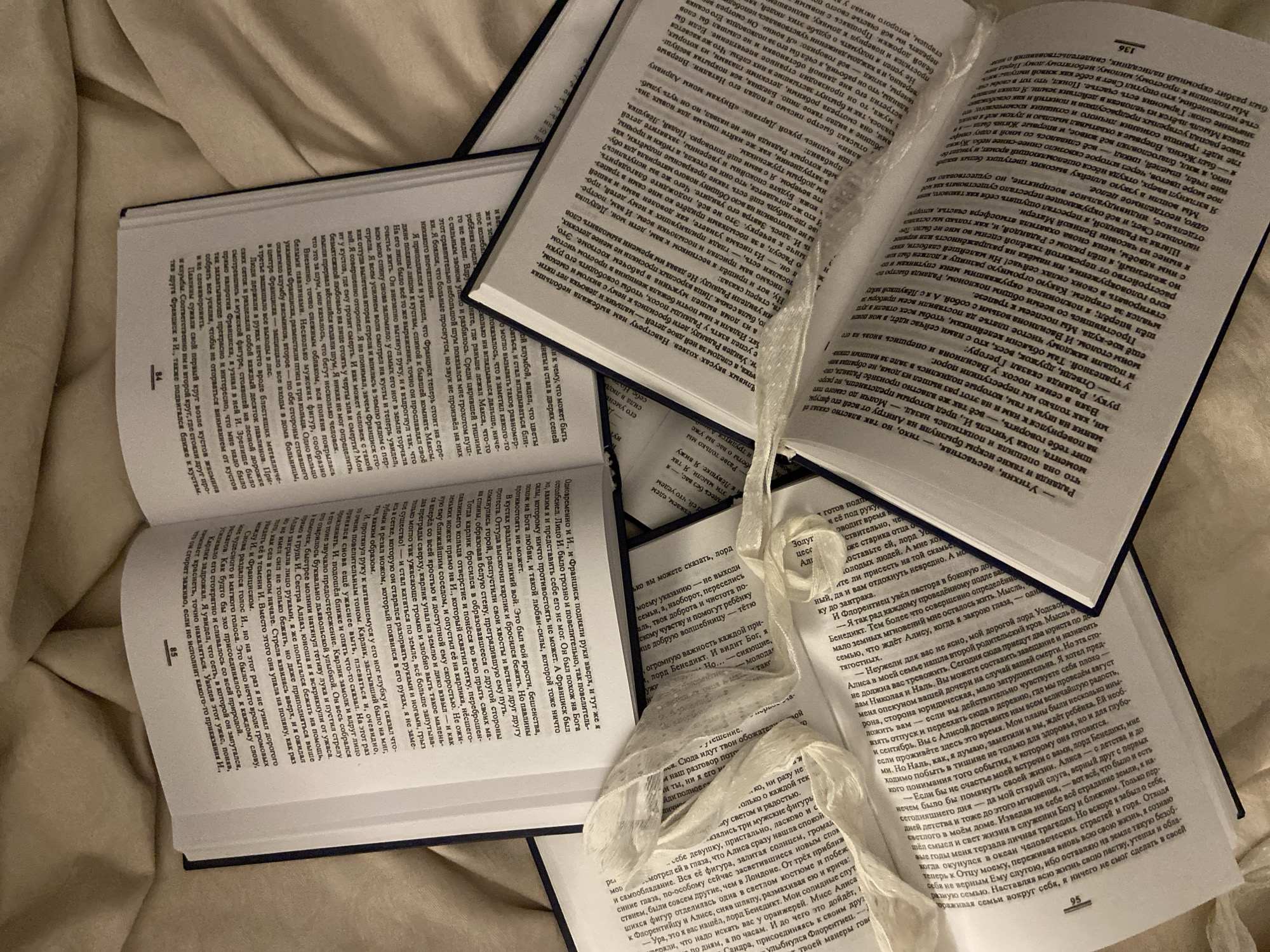 The scene depicts at least six hardcover books, open to various pages, scattered across a light-colored, soft, and textured textile surface, consistent with bedding. The visible book covers are dark blue or black, and the pages are off-white with text printed in Cyrillic script, identified as the Russian language. Specific page numbers, including 84, 85, 95, and 136, are visible. A thin, white, woven ribbon or string is intertwined among several of the lower books. No individuals are present. The precise location within the city of Budva, Montenegro, cannot be determined from the visual information provided.