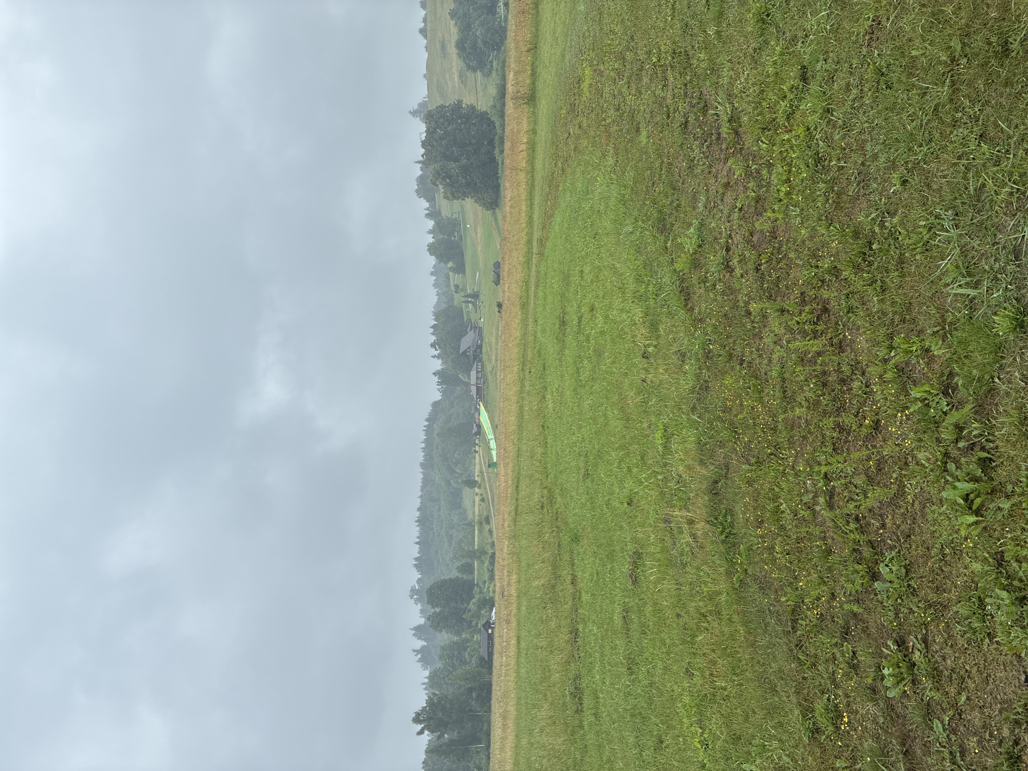 The image depicts a cloudy, overcast day in the city of Sabile, Latvia. In the foreground, there is a wide-open field of green grass with some patches of yellow wildflowers. The grass appears somewhat uneven and mowed. In the middle ground, there is a dirt path or road leading towards a cluster of buildings and trees. One building appears to be a dark-colored house, with a white car parked in front of it. Further to the right, there is a bright green structure, possibly a ramp or slide, set against a backdrop of trees and rolling hills. The background consists of a dense forest of green trees covering the hills, under a heavy, gray sky. The overall impression is of a rural or semi-rural landscape on a day with poor weather.