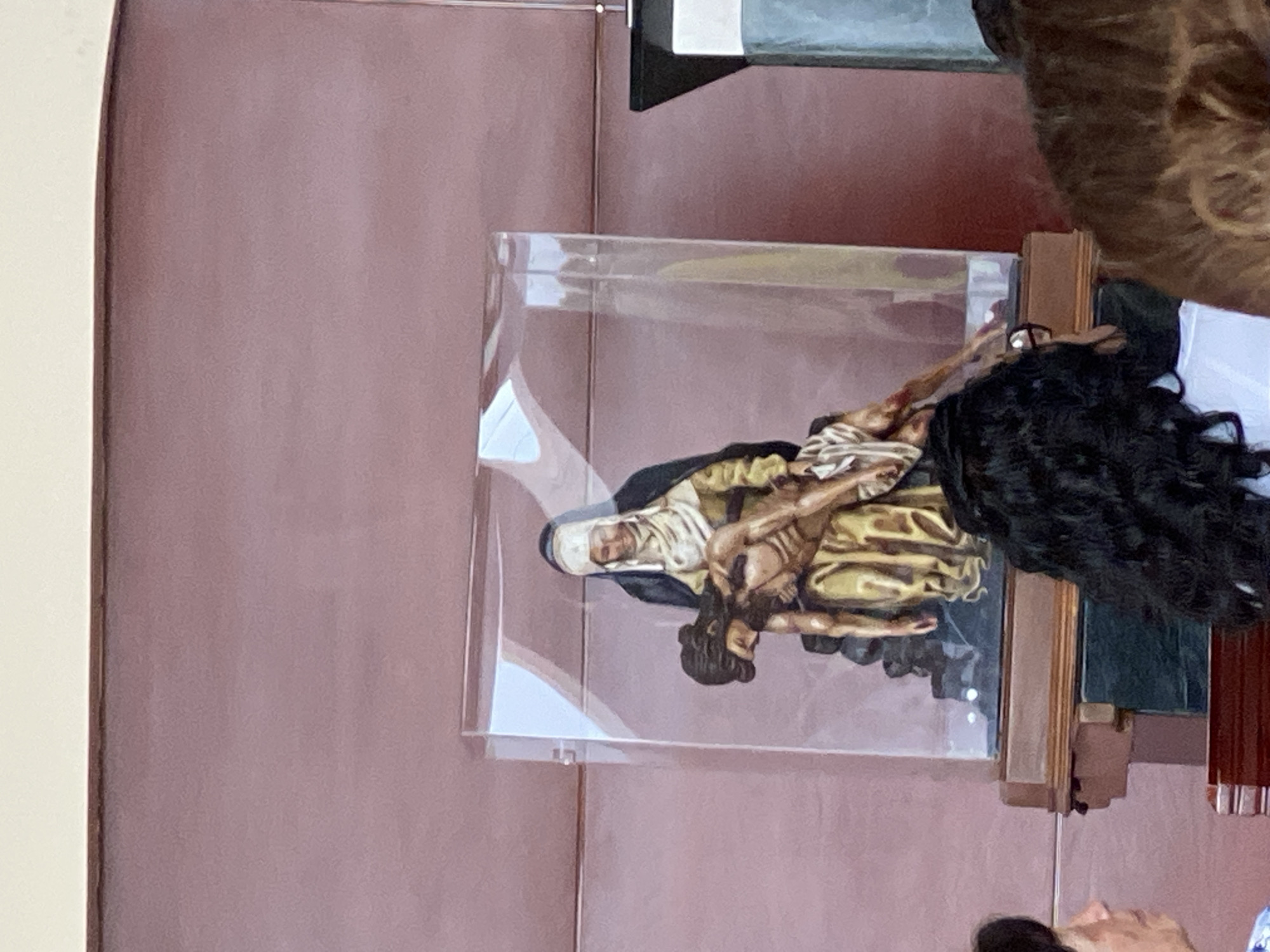 The image shows a depiction of the Pietà encased in a clear, possibly acrylic, box. The sculpture features Mary holding the body of Jesus. The sculpture is displayed indoors against a mauve-colored wall. Part of a person with curly black hair is visible in the foreground, suggesting they are observing the sculpture. Other people are visible to the lower right. The location is likely in San Bernardino Tlaxcalancingo, Mexico.