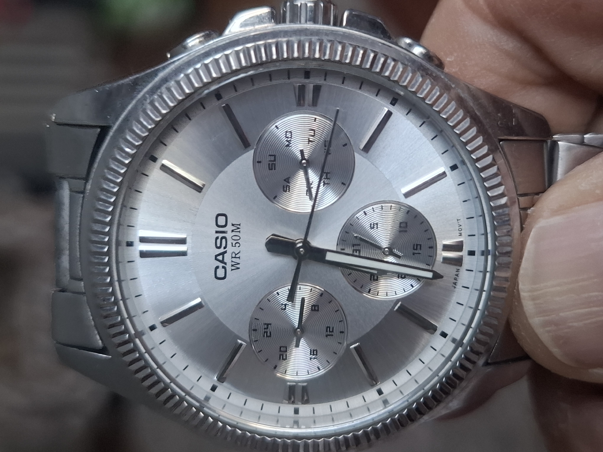 A silver Casio watch with a textured dial is displayed. The watch features three subdials: one at the 3 o'clock position showing days of the week (SU, MO, TU, TH, SA indicated), another at the 9 o'clock position with numbers (12, 16, 20, 24 indicated), and a third at the 6 o'clock position with numbers (10, 15, 31 indicated). The watch has silver hands and hour markers, and the brand name "CASIO" and "WR 50M" are visible on the dial. The watch band is also made of silver-colored metal. The image is a close-up of the watch, with a portion of a person's finger visible in the lower part of the frame. The background is blurred and out of focus. The image does not contain any information that would allow for the identification of its location in the city of Almaty, Kazakhstan.