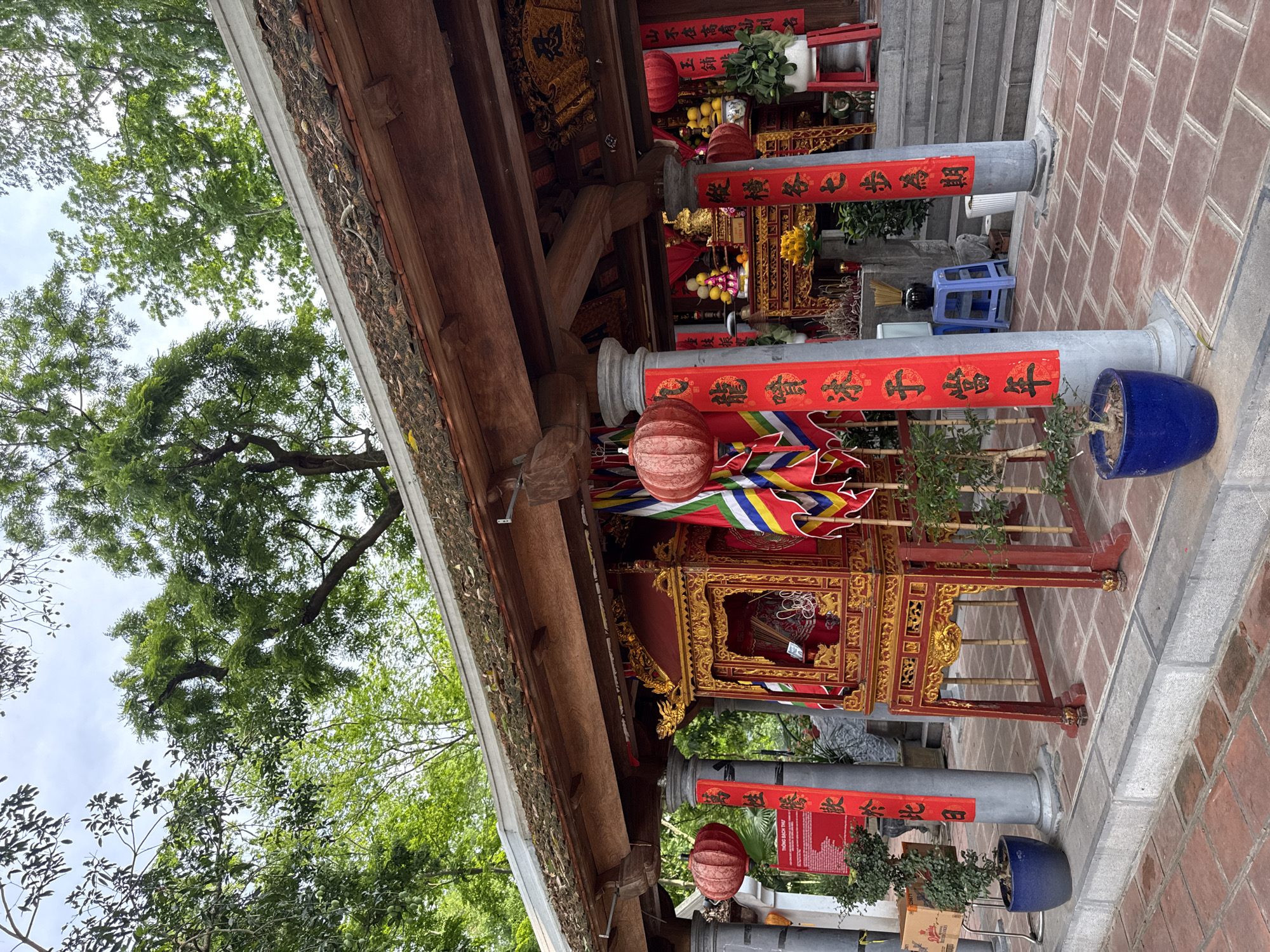 The image depicts a temple structure in Hà Nội, Vietnam. The building is adorned with red and gold decorations and colorful banners with vertical black calligraphy. Gray pillars are wrapped with red banners that also have black lettering. Lanterns hang from the roof. The roof is traditional Vietnamese style with gray brick roof tiles. The temple is set on a brick courtyard. Green trees surround the temple. Parts of the roof and the sky are visible.
