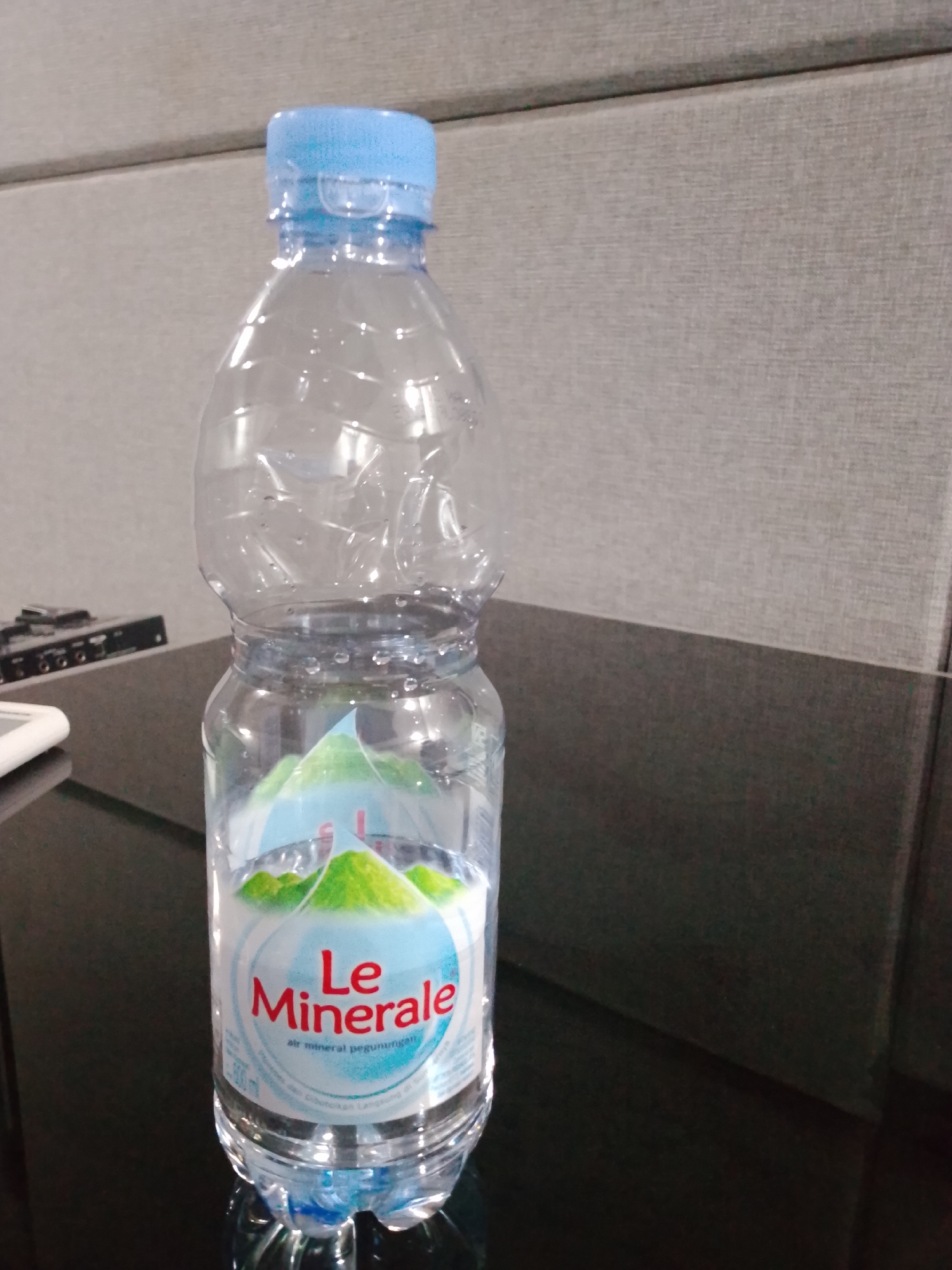 A clear plastic bottle of Le Minerale mineral water stands on a dark, reflective surface, possibly a table. The bottle is partially full of water, with condensation visible on the exterior. The label on the bottle features the "Le Minerale" logo in red text, with an image of green mountains against a light blue background. Text on the label indicates "air mineral pegunungan" (mountain mineral water). The background consists of a textured, light grey surface and a darker grey surface below it. To the left of the bottle, a white rectangular object, possibly a phone or tablet, and a black electronic device with buttons and ports are partially visible. The lighting is from above, creating reflections on the bottle and the surface. The scene appears to be indoors, in a modern setting. The location is South Jakarta, Indonesia.