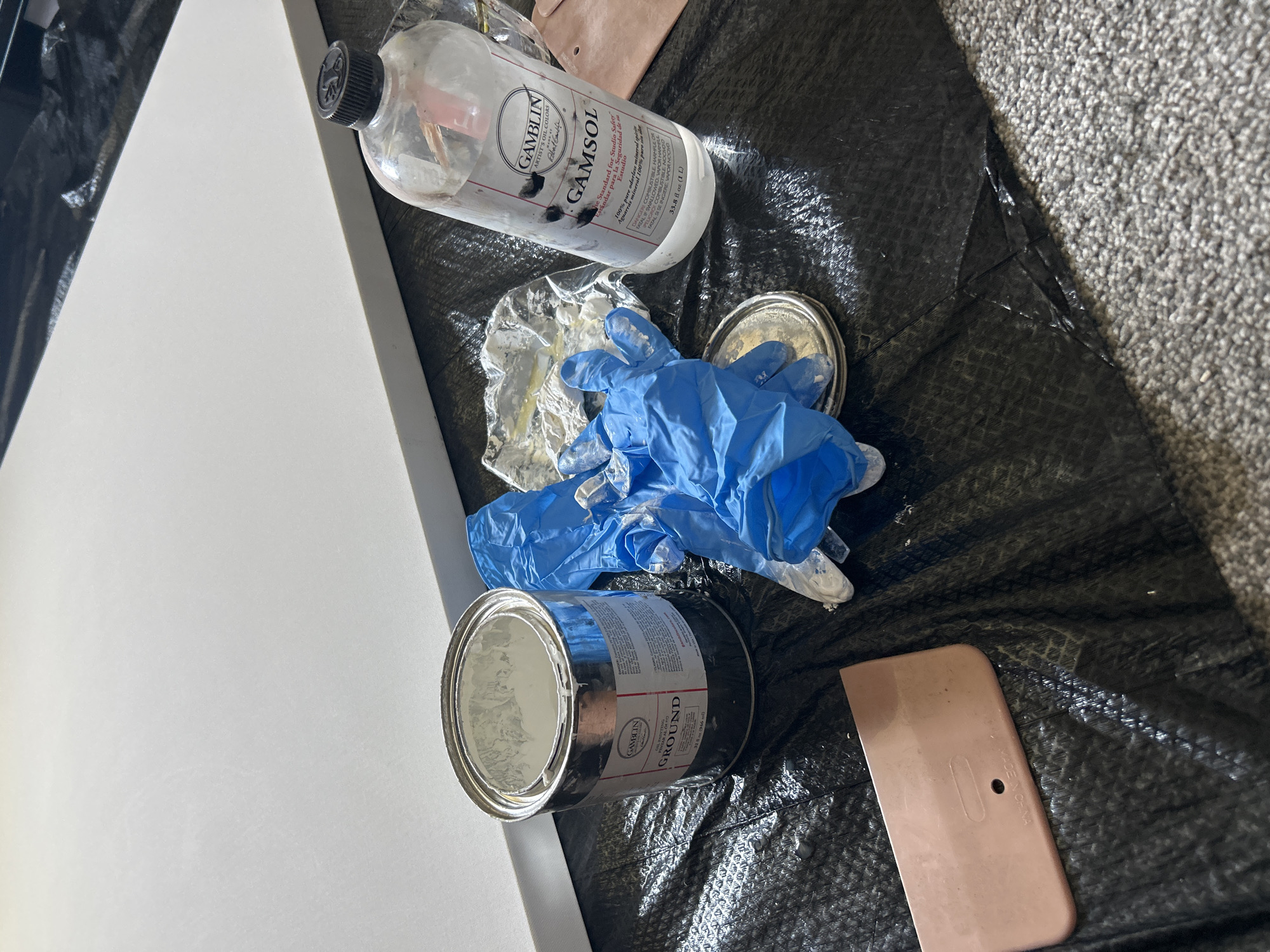 The image depicts an art studio setup, likely indoors, with various art supplies scattered on a black plastic sheet. These supplies include a canvas, a bottle of Gamblin Gamsol, an open can of Gamblin Ground, used blue latex gloves, a plastic paint scraper, and some plastic film. The plastic sheet appears to be laid out on a carpet. The text on the Gamsol bottle is in both English and Spanish, indicating it is sold in both markets. The text on the can of ground also gives the brand and type of supply as well as the measurement in fluid ounces (US). There is no clear indication of the city of Cape Coral, Florida, or the presence of any people in the image.