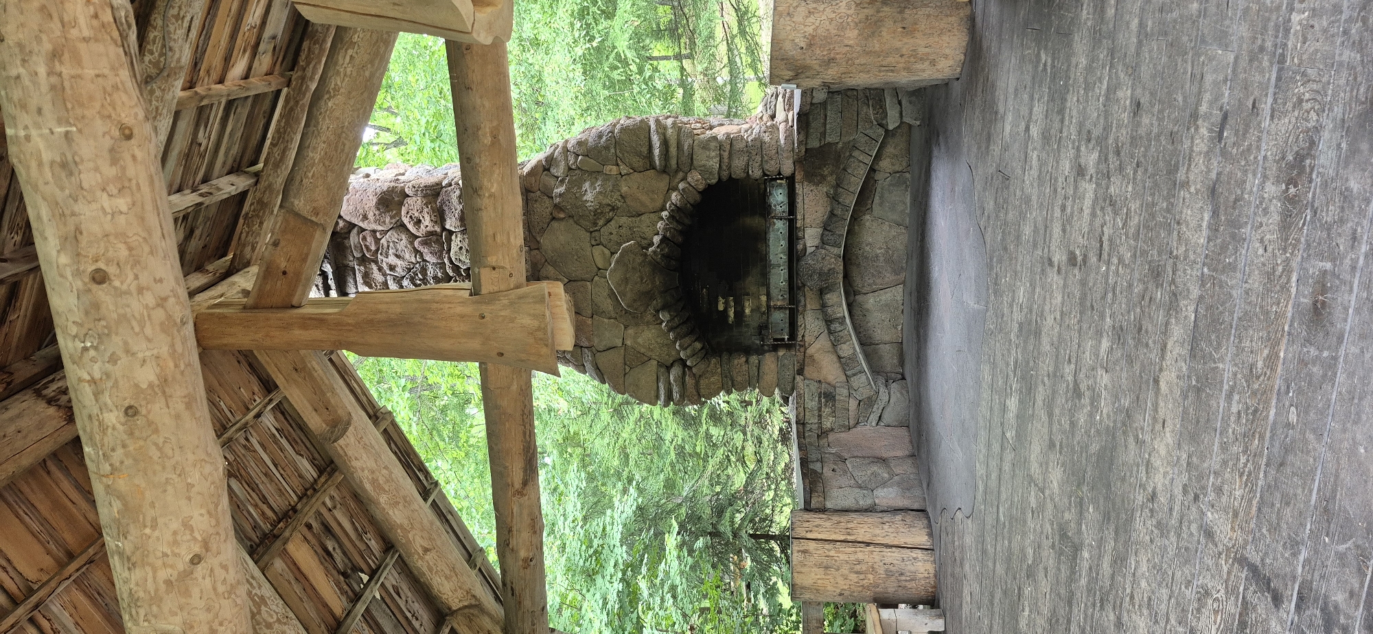This is a medium shot, vertical, interior view of a rustic outdoor pavilion with a stone barbecue. The pavilion has a roof constructed of wooden beams and planks, with exposed rafters. A large stone chimney and barbecue grill dominate the center of the image. The grill appears to be made of metal. To the left and right of the barbecue are large, rough-hewn wooden logs. The floor of the pavilion is made of weathered wooden planks. In the background, a dense forest of green trees is visible. The scene appears to be located outdoors in a natural setting, possibly a park or campsite, and there are no people present. The image does not contain information that would indicate its location in Knevichi, Russia.