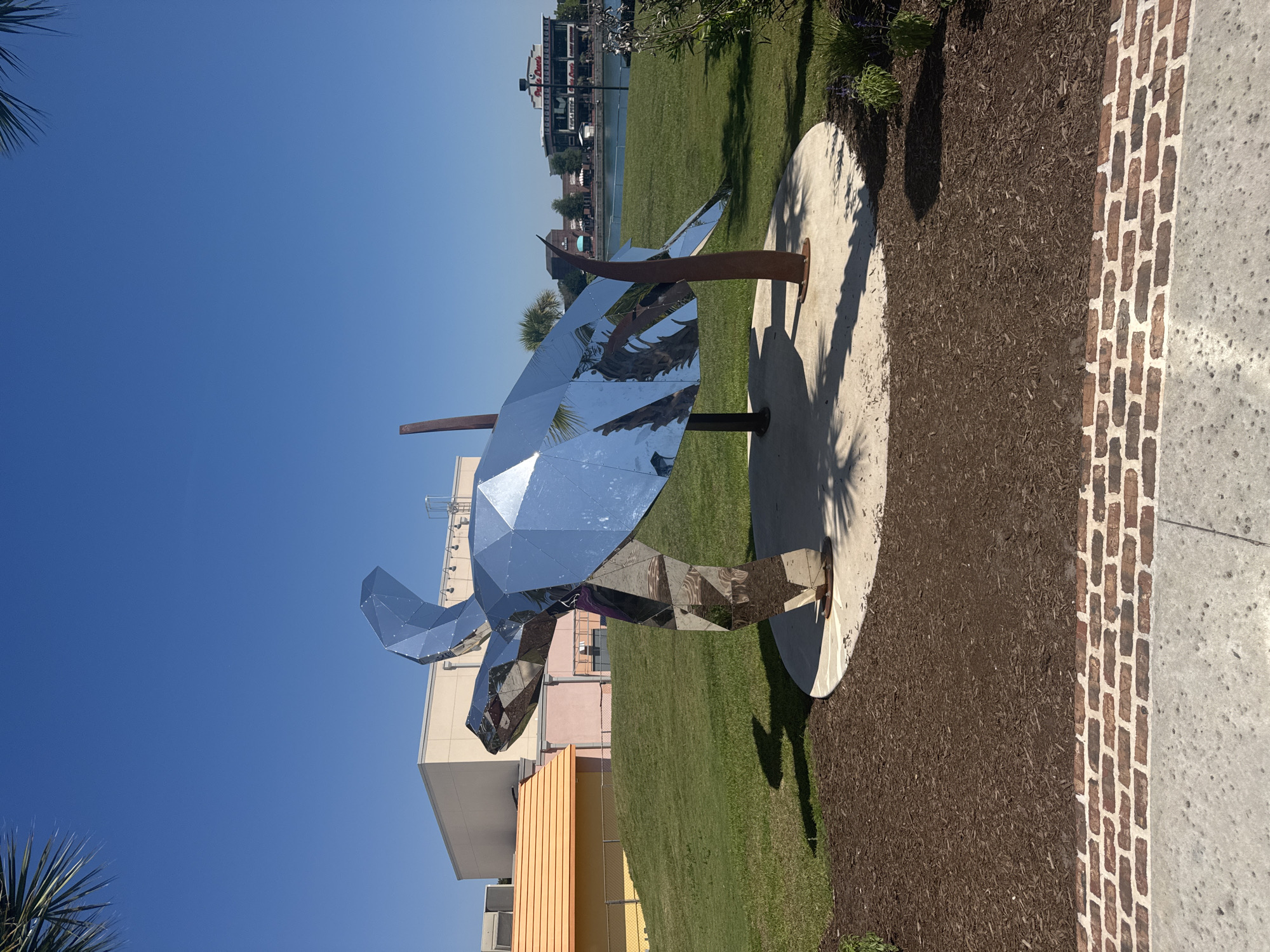 The image presents a large, faceted sculpture of a sea turtle, constructed from a highly reflective, silver-toned material, under a clear, cloudless blue sky. The sculpture stands on a circular concrete pad, surrounded by dark brown mulch, which transitions to a manicured green lawn. Two dark brown, organically shaped posts provide additional support to the turtle. The reflective surfaces of the sculpture mirror segments of a palm tree, the blue sky, and other elements of the environment.
Distinct shadows are cast by the sculpture and its supports onto the concrete base and mulch, indicating bright sunlight. In the midground, to the left of the sculpture, a grassy hill ascends towards a light-colored building with a flat roof, and an adjacent structure featuring a bright orange, sloped roof. A chain-link fence runs along the base of the hill.
To the right of the sculpture, a body of water is visible, beyond which lies a commercial complex. Prominently displayed on one of the buildings across the water is a sign reading "Paula Deen's Family Kitchen," with other retail or restaurant establishments visible nearby. Sparse green foliage is present in the lower right corner of the foreground.
The immediate foreground consists of a textured concrete path, bordered by a strip of reddish-brown brickwork, which abuts the mulched area. Portions of palm fronds are visible in the extreme upper left and upper right corners of the frame.
The presence of "Paula Deen's Family Kitchen" and the distinctive commercial waterfront development identify the location as Broadway at the Beach in Myrtle Beach, South Carolina, United States of America. No individuals are present in the depicted scene.