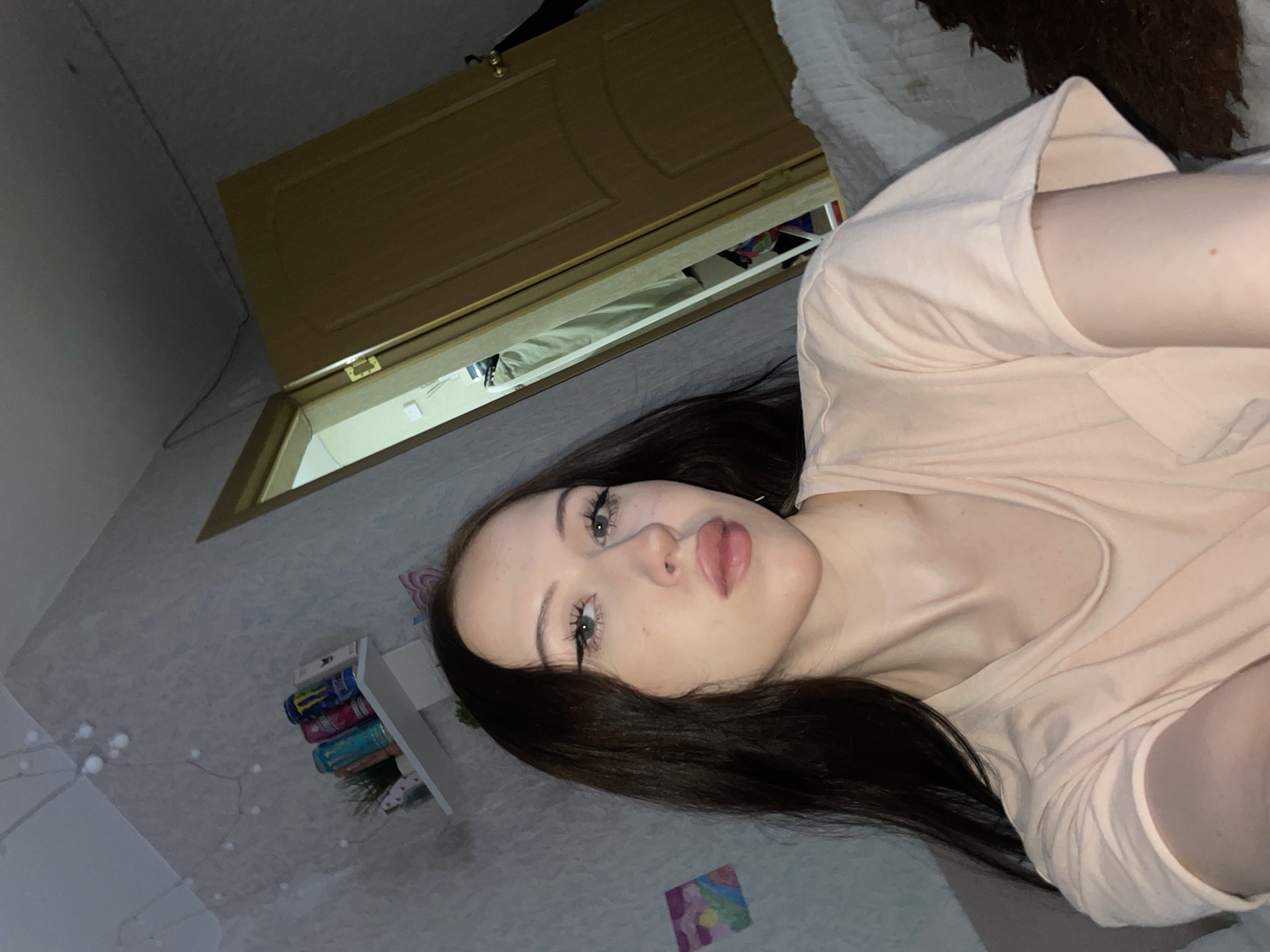 The image is a selfie taken indoors. A young Caucasian woman with long dark hair and light skin is the primary subject, positioned in the foreground and looking towards the camera. She is wearing a light peach-colored t-shirt. Her makeup includes winged eyeliner and lip gloss. In the background, a light-colored wall with a textured pattern is visible. A white shelf is mounted on the wall, displaying several cans, including what appear to be energy drink cans, and a small potted plant. To the right of the woman and further in the background, a wooden door is ajar, revealing a glimpse of a room with clothing on hangers and what looks like a tall mirror. A string of small lights is draped along the top of the wall. The overall lighting suggests it is evening or indoors with artificial light. The location is identified as Votkinsk, Russia, implying the setting is likely a residential room within the city.