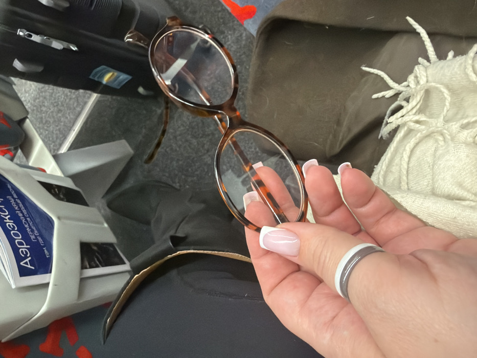 The scene is depicted within a passenger transit vehicle, specifically an Aeroexpress train, in the city of Ям, Russia. A person's left hand, with light skin and manicured fingernails featuring a French tip design, holds a pair of tortoiseshell-patterned eyeglasses with clear, oval-shaped lenses. A two-banded ring, white over gray, is worn on the thumb of the holding hand. Below the hand, a light beige, fringed scarf or blanket is partially visible, next to a section of dark olive green or brown fabric, possibly a jacket or coat. To the left, the lower leg of an individual wearing dark trousers and a black shoe or boot is visible. Above this, a white plastic seat-back pocket contains a leaflet with "Аэроэкспресс" (Aeroexpress) prominently displayed in blue Cyrillic text, with "ПЕРМСКИЙ КРАЙ" (Perm Krai) visible below it. The floor is covered with a dark gray, short-pile carpet. To the right, a black roll-aboard suitcase with visible handle hardware and faint blue and yellow stickers stands upright. A section of red and black patterned upholstery is partially visible on the far right.