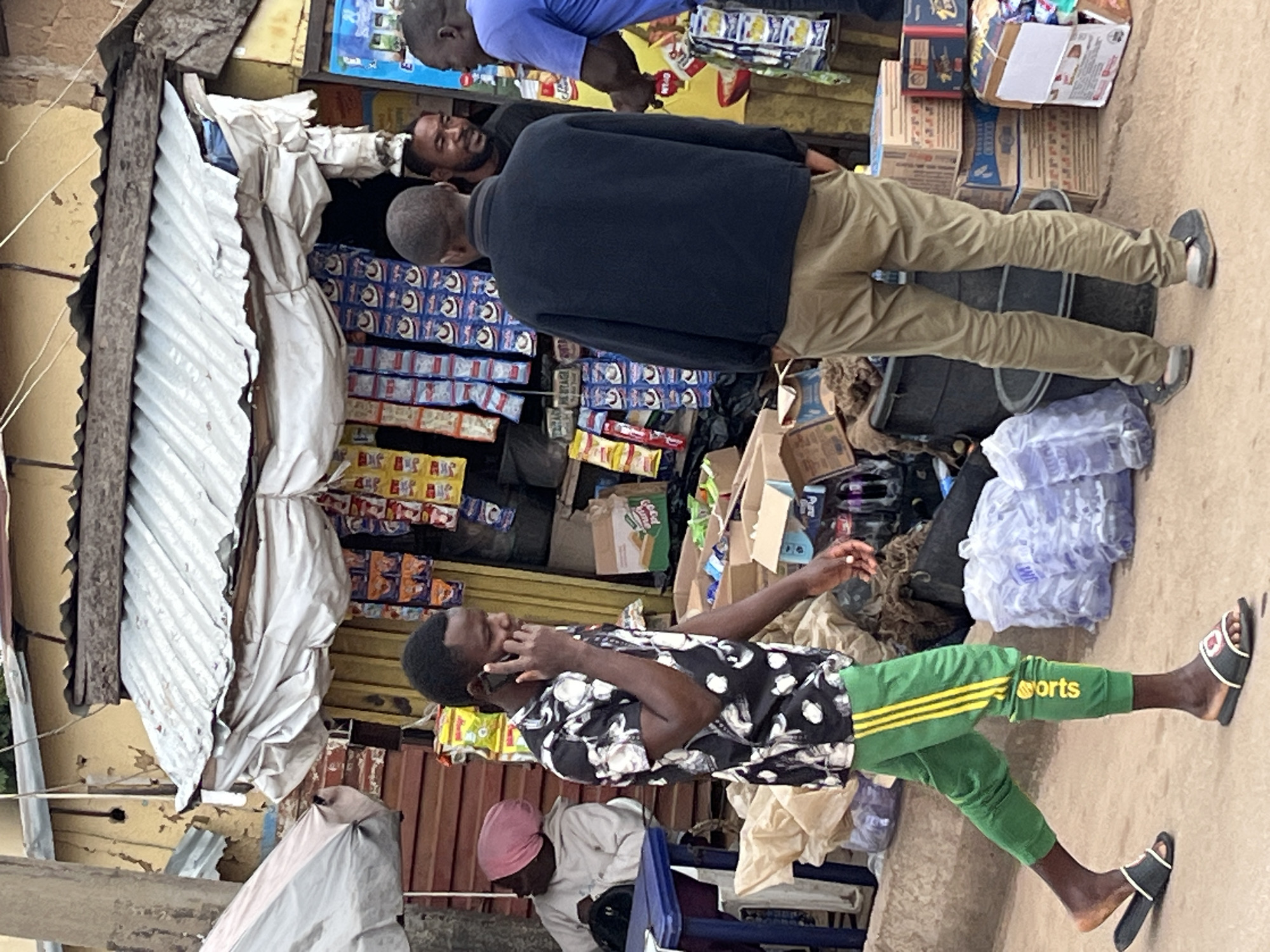 The image captures a street scene in Gura Top, Nigeria. In the foreground, a young man in a black floral shirt and green track pants with yellow stripes is walking and speaking on a mobile phone. He is wearing dark slides. Behind him, several other individuals are visible. To the left, a person wearing a pink headscarf and a light-colored garment is seated. In the center, another man in a dark jacket and khaki pants stands with his back to the camera. To the right, a man in a blue shirt is engaged in a conversation.
The environment appears to be a market or roadside stall area. Several shelves are stocked with packaged goods, including snacks and drinks. Cardboard boxes containing various items are stacked on the ground, and large black plastic containers are present. The stall itself has a corrugated metal roof with a white fabric awning. Bags of what appear to be bottled water are also visible. The ground is paved with dirt and some concrete. The overall impression is that of a busy, informal commercial setting.