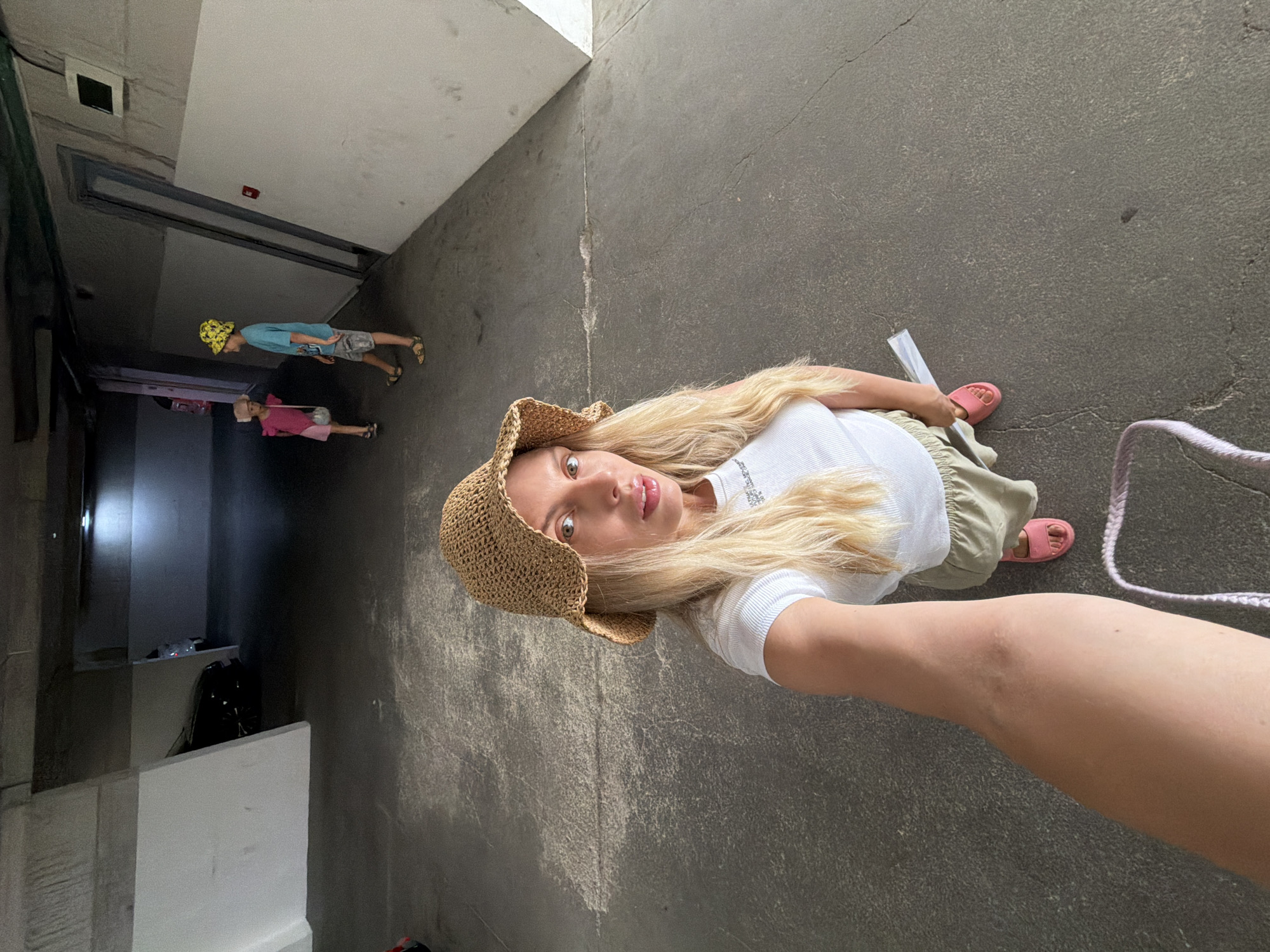 A woman in a straw hat and white top is taking a selfie in what appears to be an underground parking garage. She is wearing a light-colored skirt and pink sandals. In the background, two children are visible. One child in a pink outfit is facing away from the camera, while another child in a blue shirt and yellow hat is facing the camera. The environment is dimly lit, with concrete floors and walls, suggesting an urban, possibly residential, setting. The presence of cars in the background reinforces the parking garage context. The overall impression is a casual snapshot taken during an outing with children. The location is identified as Budva, Montenegro.