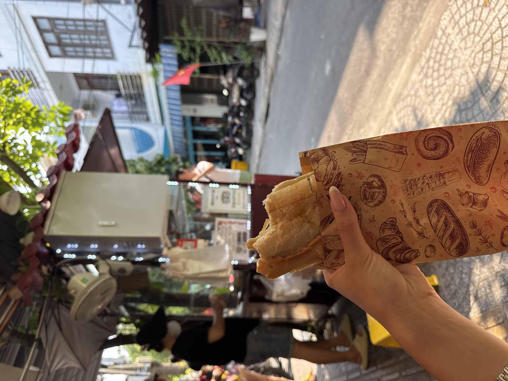 A person holds a partially eaten banh mi sandwich in a paper bag with bakery-themed illustrations. The background is a street scene with a vendor's cart, motorbikes parked along the street, and buildings. The location is likely in Đà Nẵng, Vietnam, given the food item and the architectural style of the buildings. The vendor is visible near the cart.