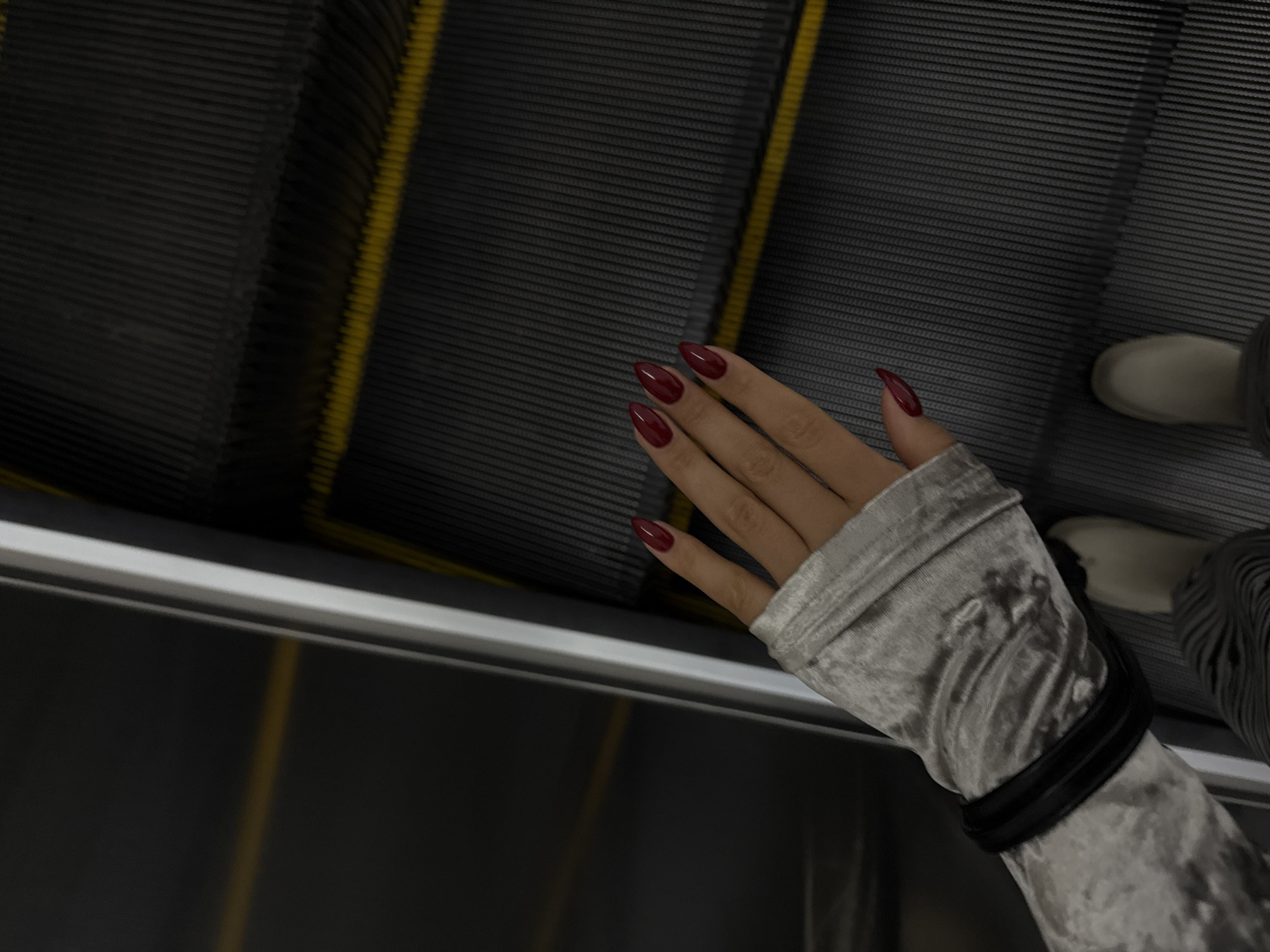 The image shows a hand with long, burgundy-colored nails and a gray, velvet-like sleeve with a black band at the wrist, resting on the moving steps of an escalator. The escalator steps are ribbed with yellow delineators. The image suggests an indoor location, possibly a shopping mall or public transportation hub. The presence of the escalator is indicative of a building with multiple levels. The exact location in Yerevan, Armenia is not discernible from this single image. Other people and their actions are not seen, only a foot wearing a light-colored shoe is partially visible on the escalator step behind the hand.