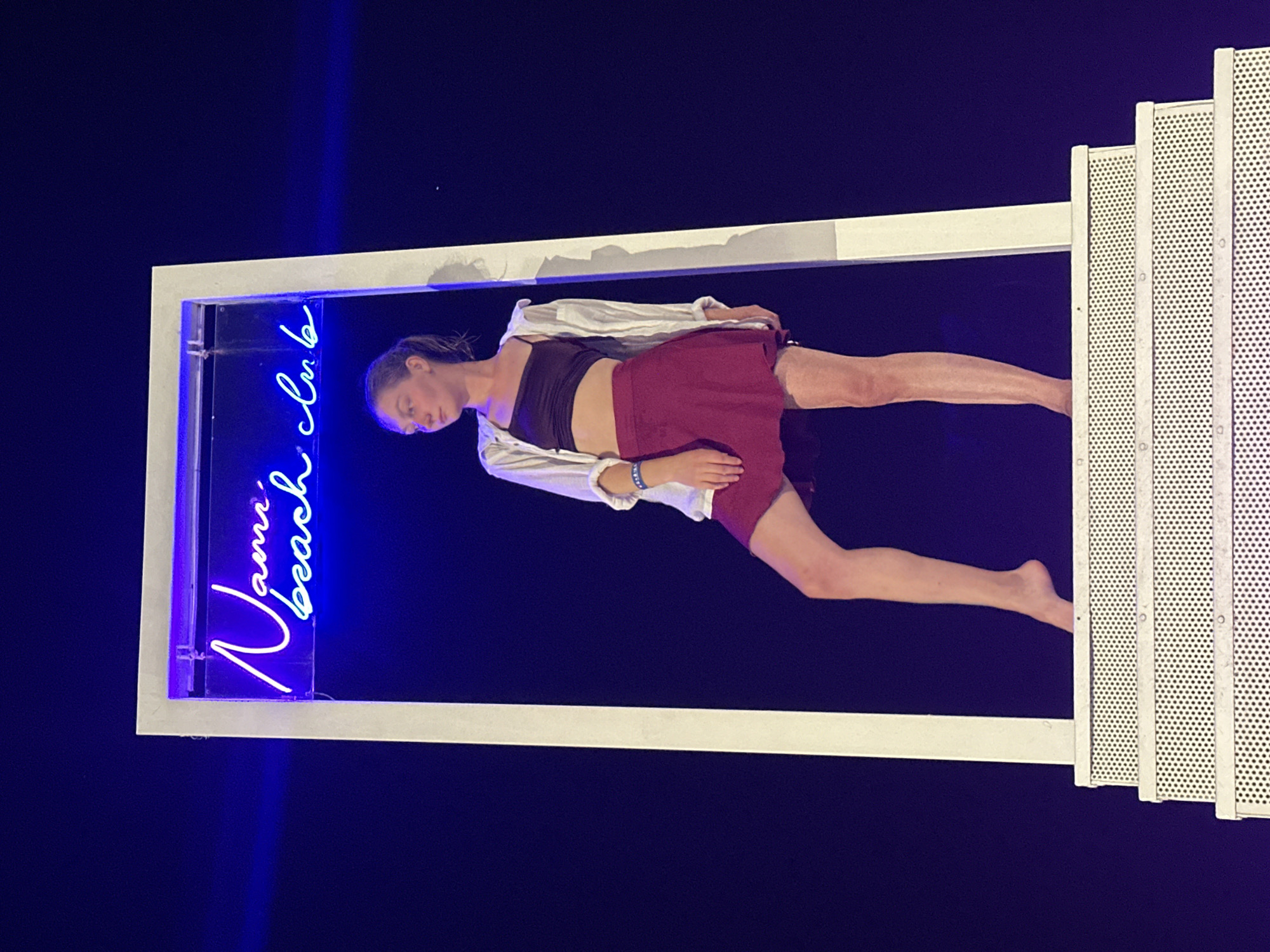 This media file captures a stylized scene featuring a young woman in what appears to be a performance or artistic presentation. She is positioned within a rectangular frame, illuminated by a bright blue neon sign that spells out "Nami beach club" in cursive script. The woman is dressed in a dark tank top, a light-colored open shirt, and a maroon skirt. Her body is contorted, with one leg extended and her arm resting on her skirt. She has a pensive or serene expression, with her eyes closed.
The background is a deep, dark blue, suggesting nighttime or a darkened studio. To the right of the frame, a white, perforated structure, possibly a set piece or part of a stage design, is visible, adding a contrasting element to the dark background and neon lighting. The overall atmosphere is dramatic and contemporary, likely set at an event or performance in Đà Nẵng, Vietnam, as indicated by the location context. The neon sign is a prominent visual element, directly contributing to the "beach club" theme. The image has been rotated, presenting the scene from a vertical perspective.