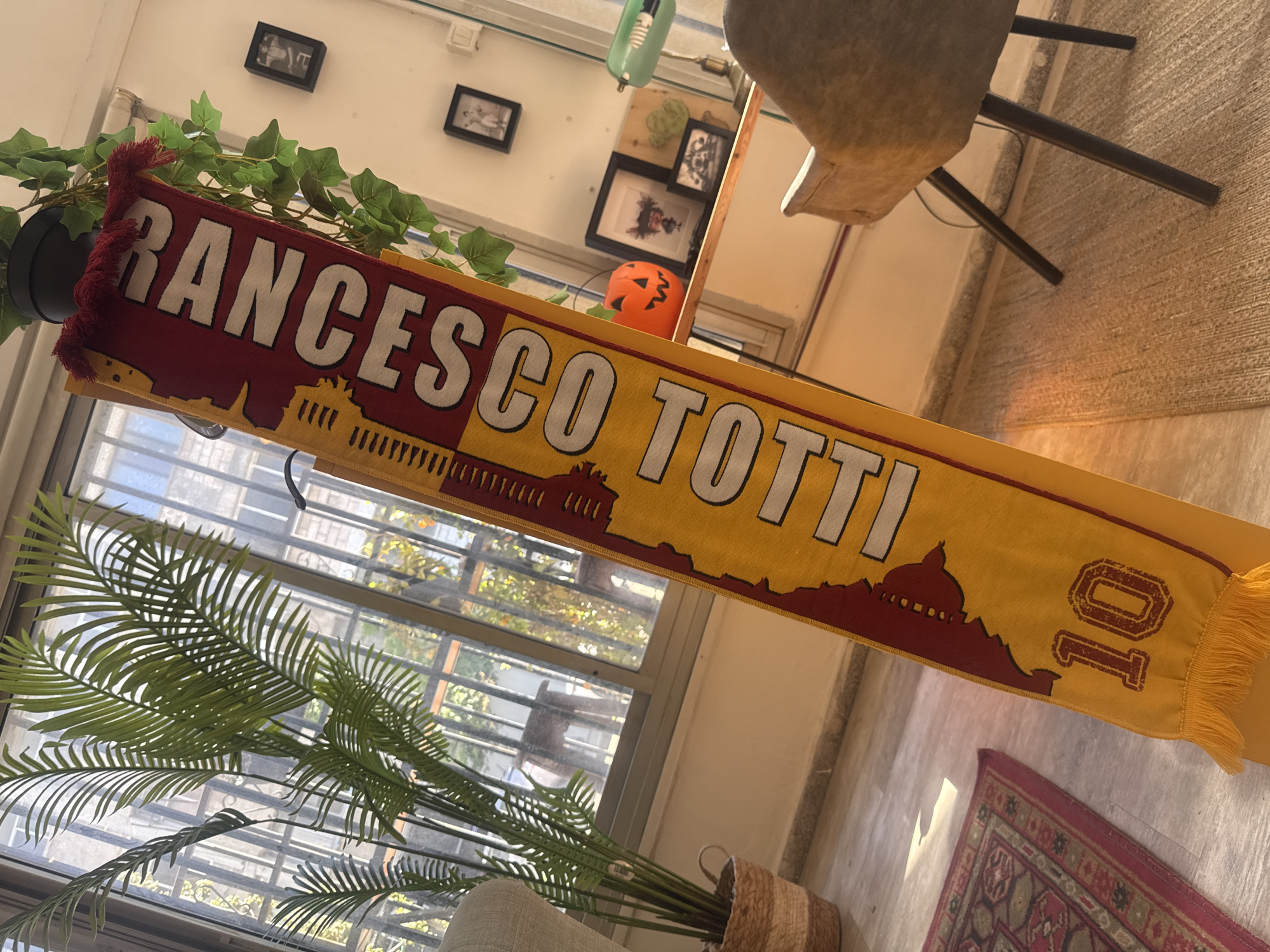 A brightly lit indoor scene features a "FRANCESCO TOTTI" scarf draped across the frame. The scarf is predominantly yellow and red with white lettering, bearing the number "10" at one end and an outline of a city skyline. Behind the scarf is a large window providing ample natural light. A potted palm plant stands in front of the window, its fronds partially obscuring the view. A smaller potted plant sits to the left. A small orange plastic pumpkin is visible behind the scarf. In the background, framed pictures hang on the wall. A chair sits to the right of the frame, partially out of view. A patterned rug sits on the floor. The location is the city of Tel-Aviv, Israel.