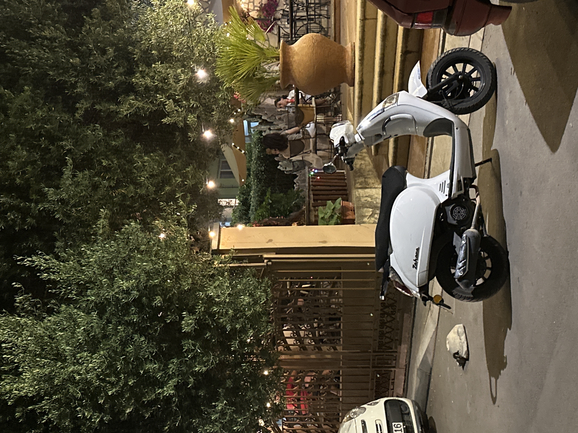 In Budva, Montenegro, a white scooter is parked on a paved street at night. The scooter is facing right and has its kickstand down. Behind the scooter, a wrought-iron fence encloses an outdoor seating area for a restaurant or cafe. Several people are seated at tables in the background, some of whom are visible through the fence. Large trees with string lights are overhead, casting a warm glow on the scene. A white car is parked to the left of the scooter, and a red car is partially visible to the right. A large terracotta pot with a palm plant stands on a set of stairs to the right of the scooter. The overall atmosphere suggests a lively evening in a Mediterranean town.
