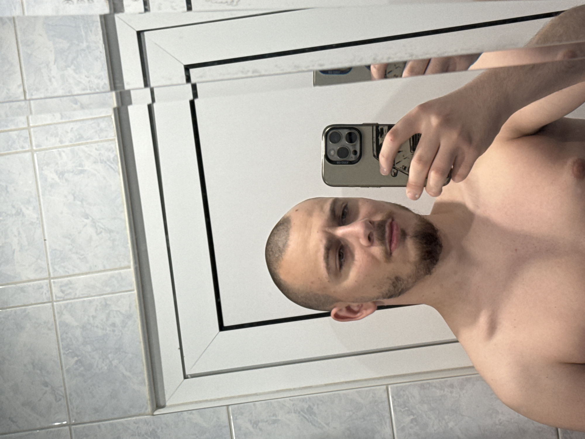 A young man is captured in a mirror selfie, shirtless and holding a smartphone with a light-colored case in his right hand. He has very short, buzzed hair and a short beard or stubble. His expression is neutral. The smartphone's camera array is visible, and the text "NO COOL" can be seen on its case.
The setting appears to be a bathroom, with light-colored, possibly light blue or grey marble-patterned tiles covering the walls to the left and bottom of the mirror. The mirror itself is framed, possibly composed of multiple rectangular panels with black dividers, creating a structured reflection. The man's arm and shoulder are visible in the reflection, along with parts of the room beyond. This scene is located in Stara Zagora, Bulgaria.