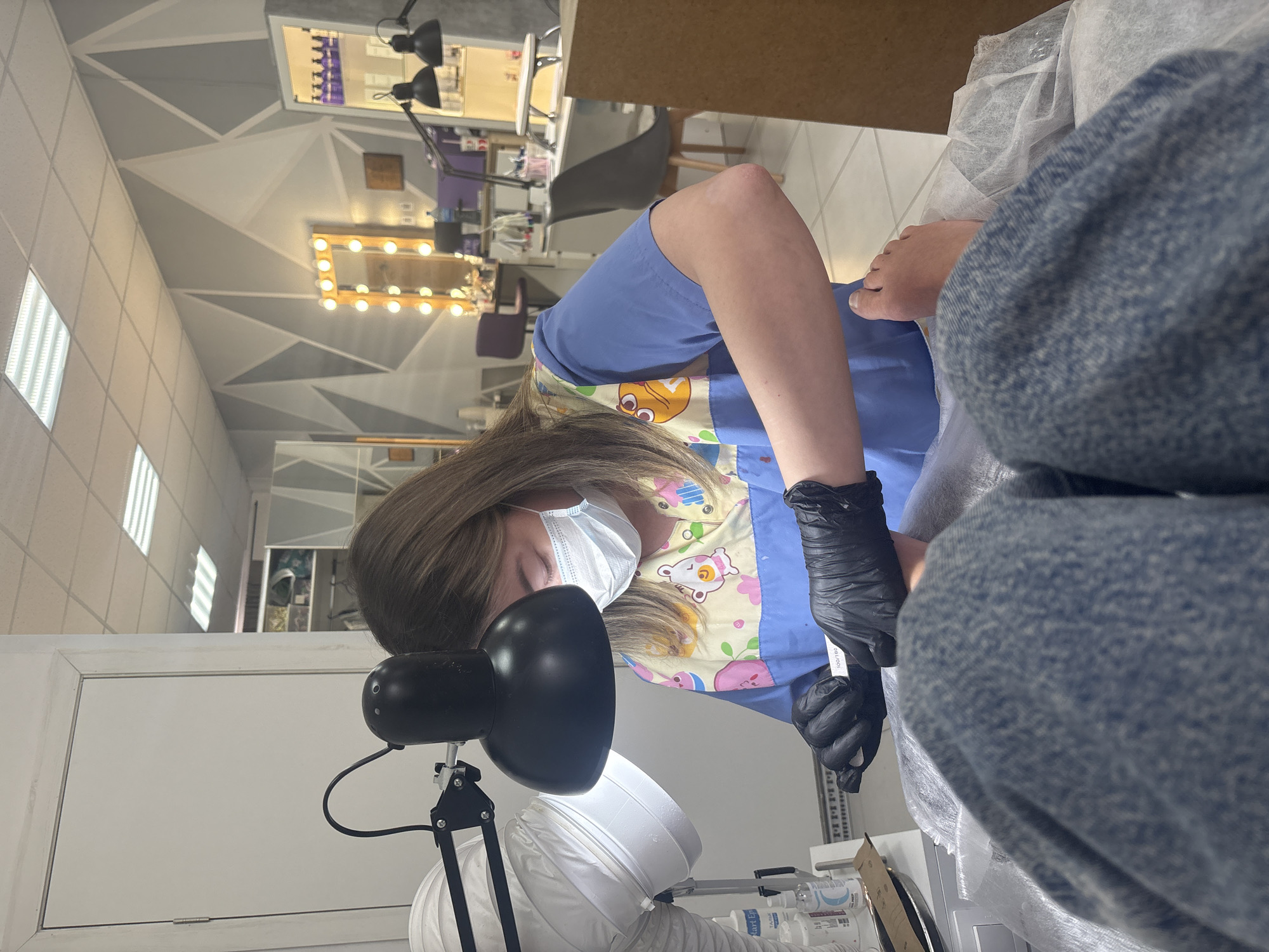 A fair-skinned woman wearing a light blue scrub top with a pastel-colored patterned trim, a white face mask, and black latex gloves is performing a pedicure on a person. The person receiving the pedicure is wearing denim jeans. A black gooseneck lamp is directed toward the pedicure area. Disinfectant supplies are arranged on a white surface next to the pedicure chair. The walls are painted in a geometrical design with gray and white paint. A lighted mirror in the background is reflected in the salon mirror. The photograph was taken indoors, with the specific location being Perm, Russia.
