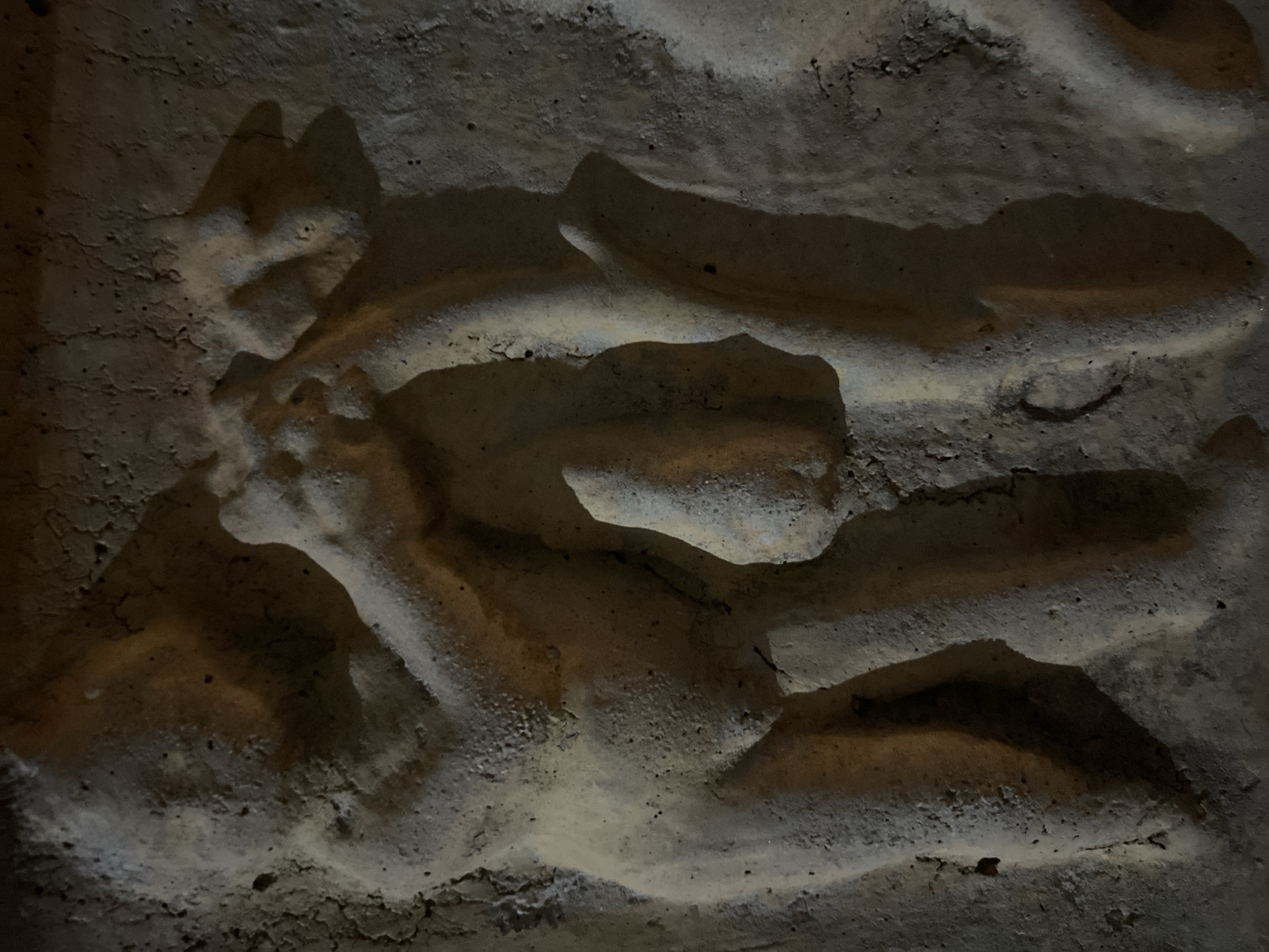 The image depicts a textured, uneven surface that appears to be a wall or rock formation. The surface is composed of a mix of light and dark shades, with shadows creating depth and highlighting the rough texture. The composition is focused on the details of the surface itself, with no clear indication of people, subjects, or objects beyond the wall or rock formation. The environment is undefined due to the close-up nature of the shot. There is no specific information within the image to confirm its location within Budva, Montenegro.