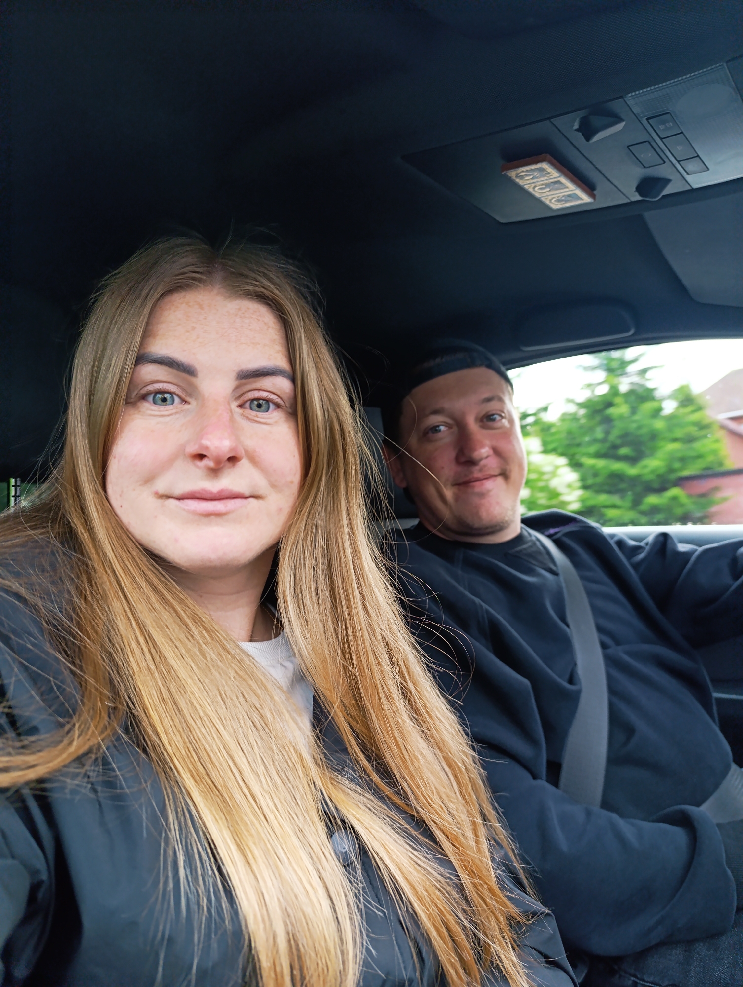 The image depicts a woman and a man seated in a car. The woman is in the foreground and slightly to the left, while the man is in the background and to the right. Both individuals are wearing dark clothing. The woman has long blonde hair, fair skin, and freckles. The man wears a black beanie. The background visible through the car window suggests an outdoor environment with greenery and a building in the distance. The car's interior is visible. The location is the city of Unknown, Unknown.