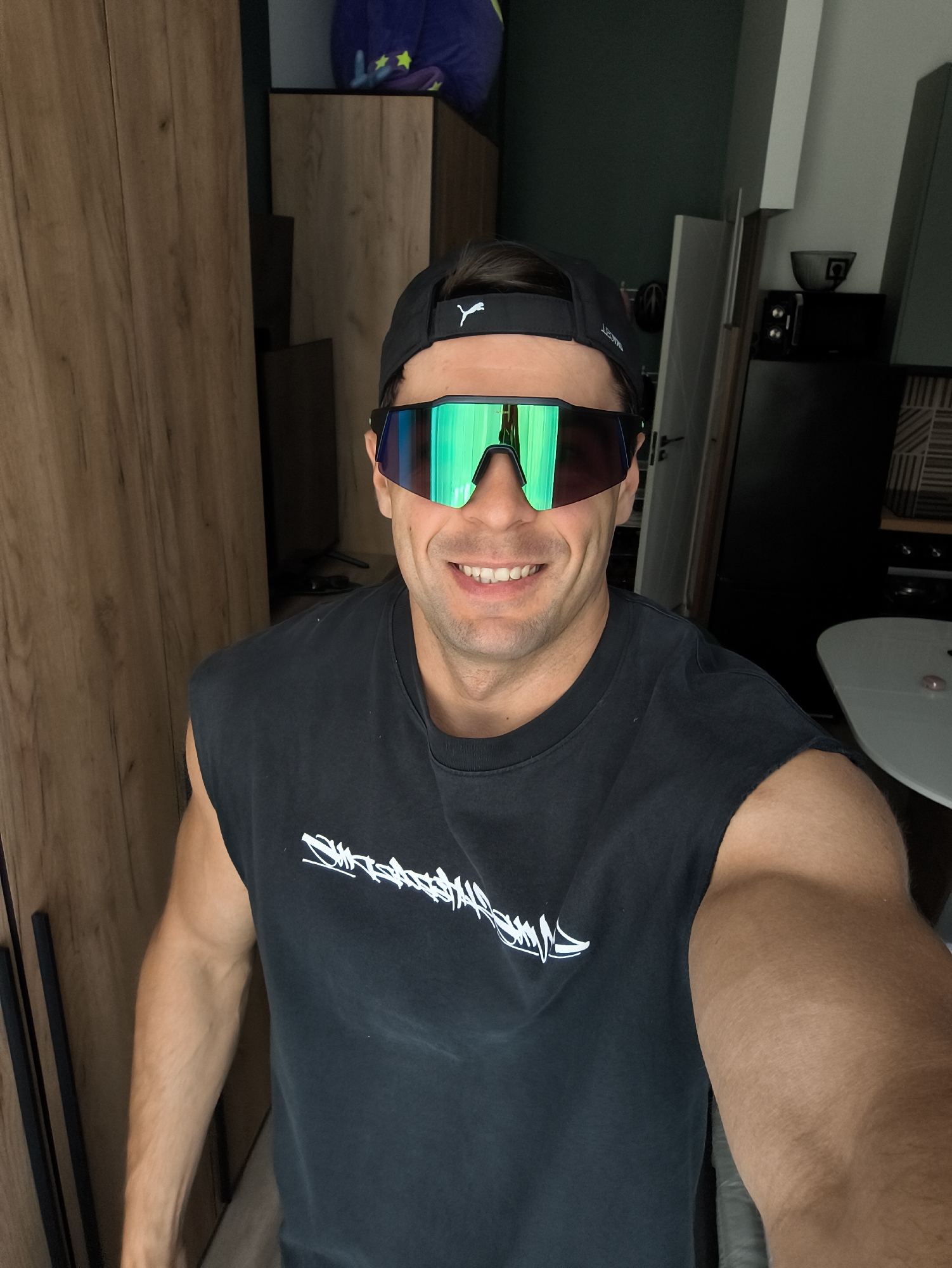 An adult male, approximately 20-40 years old, is depicted in a close-up selfie taken within an indoor residential space in Moscow, Russia. He is smiling, revealing his teeth, and has a muscular physique, with defined arms visible. He wears a black sleeveless t-shirt with a white abstract graphic design on the chest. On his head, he wears a black baseball cap backward, featuring a white Puma logo on the visible side. He is also wearing large, wraparound reflective sunglasses with vibrant green and blue mirrored lenses.
The background to the subject's left features a light-brown wooden cabinet or wardrobe with vertical panels and dark handles. To his right, the background transitions to a dark green painted wall section above a white horizontal trim. Below this, a dark countertop or surface holds a black appliance (possibly a microwave or mini-fridge) with a gray bowl placed on top. Further to the right, geometric patterned wall tiles are visible. In the far background, a white round table with a small pink translucent object rests. A purple object with yellow stars is partially visible at the top left of the background.
