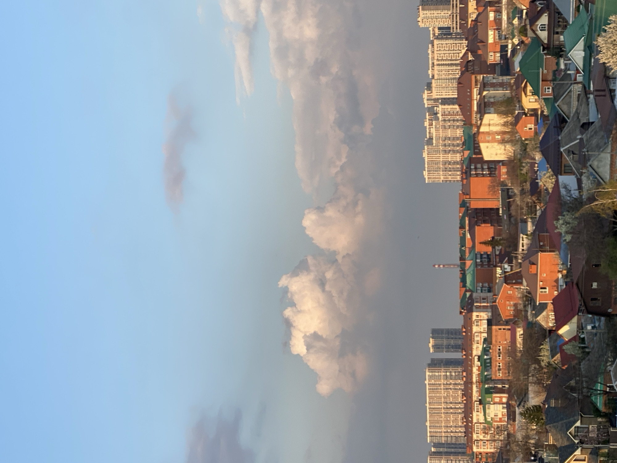 The image shows a mixed urban landscape in an unknown city. High-rise apartment buildings are interspersed with lower-density residential housing with varied roof colors (red, green, blue). A construction crane is visible amidst the buildings. The sky is partially covered with white and grey clouds against a blue backdrop. There are trees with spring blossoms intermingled with the structures.