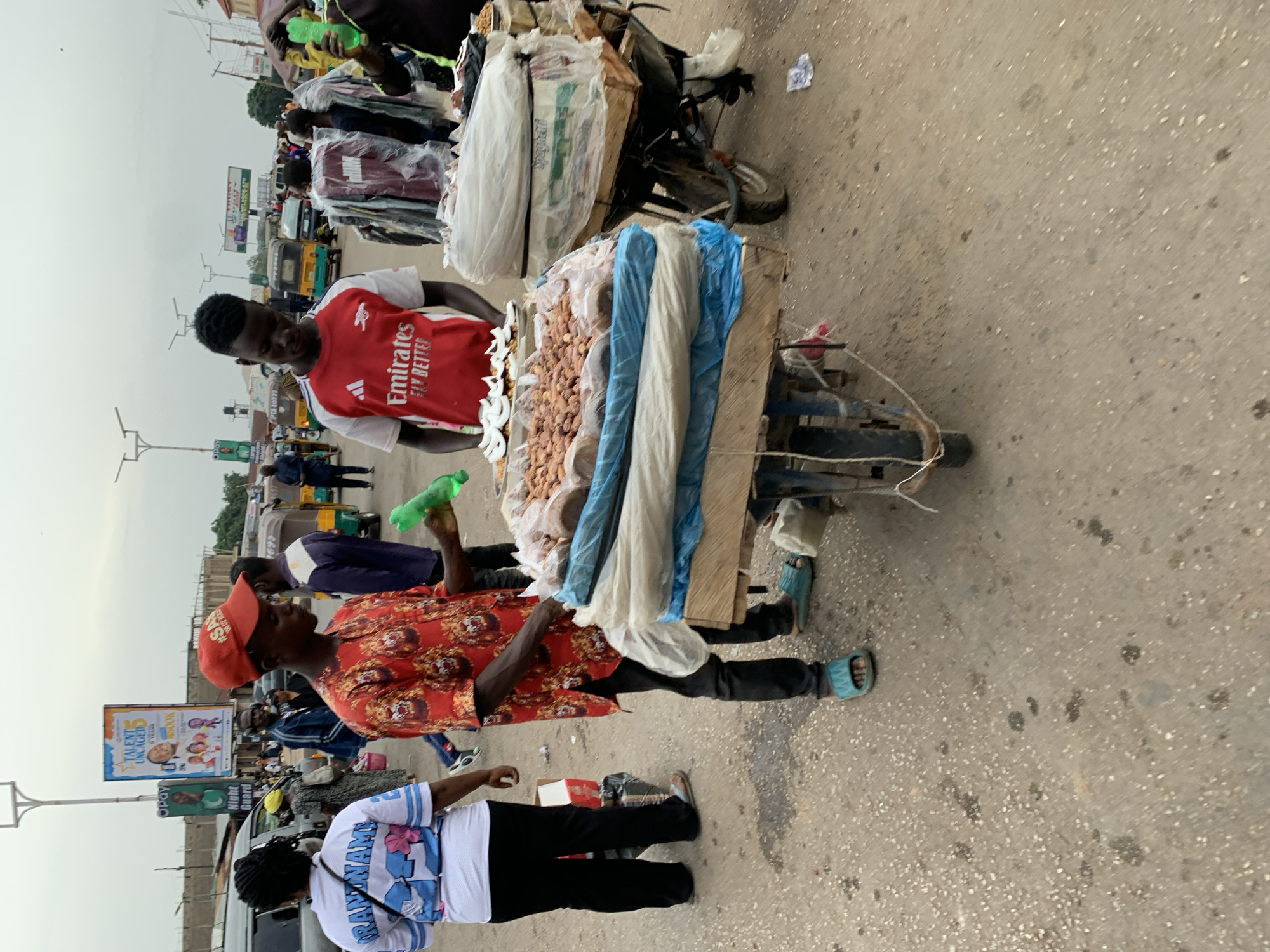 The image depicts a street scene in what appears to be Gura Top, Nigeria. A street vendor stands behind a cart laden with goods, including what appears to be peanuts. He wears a red and white soccer jersey with "Emirates FLY BETTER" printed on it. To the vendor's right, another person in a patterned red shirt and red cap holds a green bottle. A woman in a white and blue shirt with the word "RANDNAME" is also visible in the lower left corner of the image. The cart is attached to a motorcycle, and there is more merchandise piled on the front of the motorcycle. Several people and vehicles, including "Keke Napep" tricycles, are in the background. There is a billboard on the left side of the image advertising "TALENT UNCAGED" with the logo "Opay" at the bottom. The street is paved and shows signs of wear and stains. The sky is overcast.