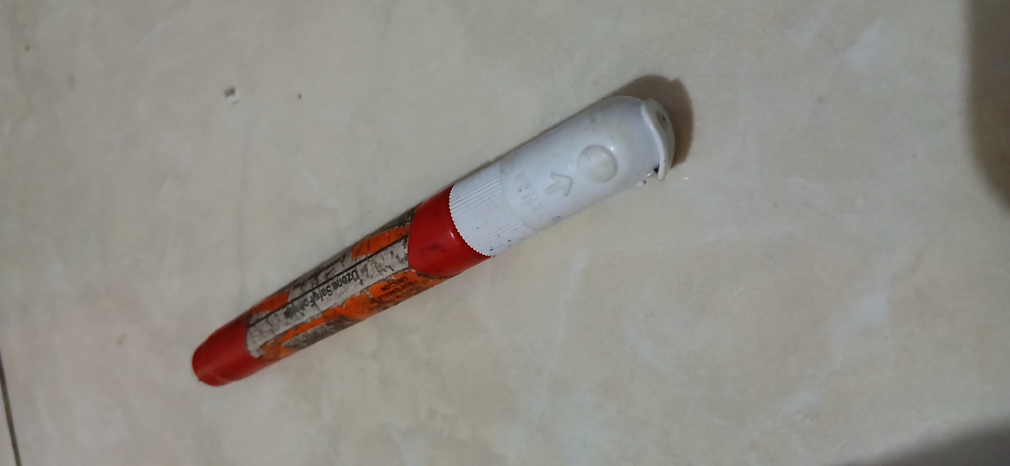 A single red and white object, resembling a pen, lies diagonally on a beige, tiled surface. The object has a red tip and a white end with a ribbed texture. A partially torn orange and white label is wrapped around the red portion of the object. The label has black text, some of which is legible and appears to be in English, with the phrase "OZONE SAFE FORMULA" visible. A faint arrow pointing left and a small circular indentation are present on the white end of the object. The background consists of a light-colored, speckled tile floor. There are no people or other discernible objects in the image. The scene appears to be indoors, given the tiled floor, and no specific geographical features are visible to indicate a location in Samarinda, Indonesia.