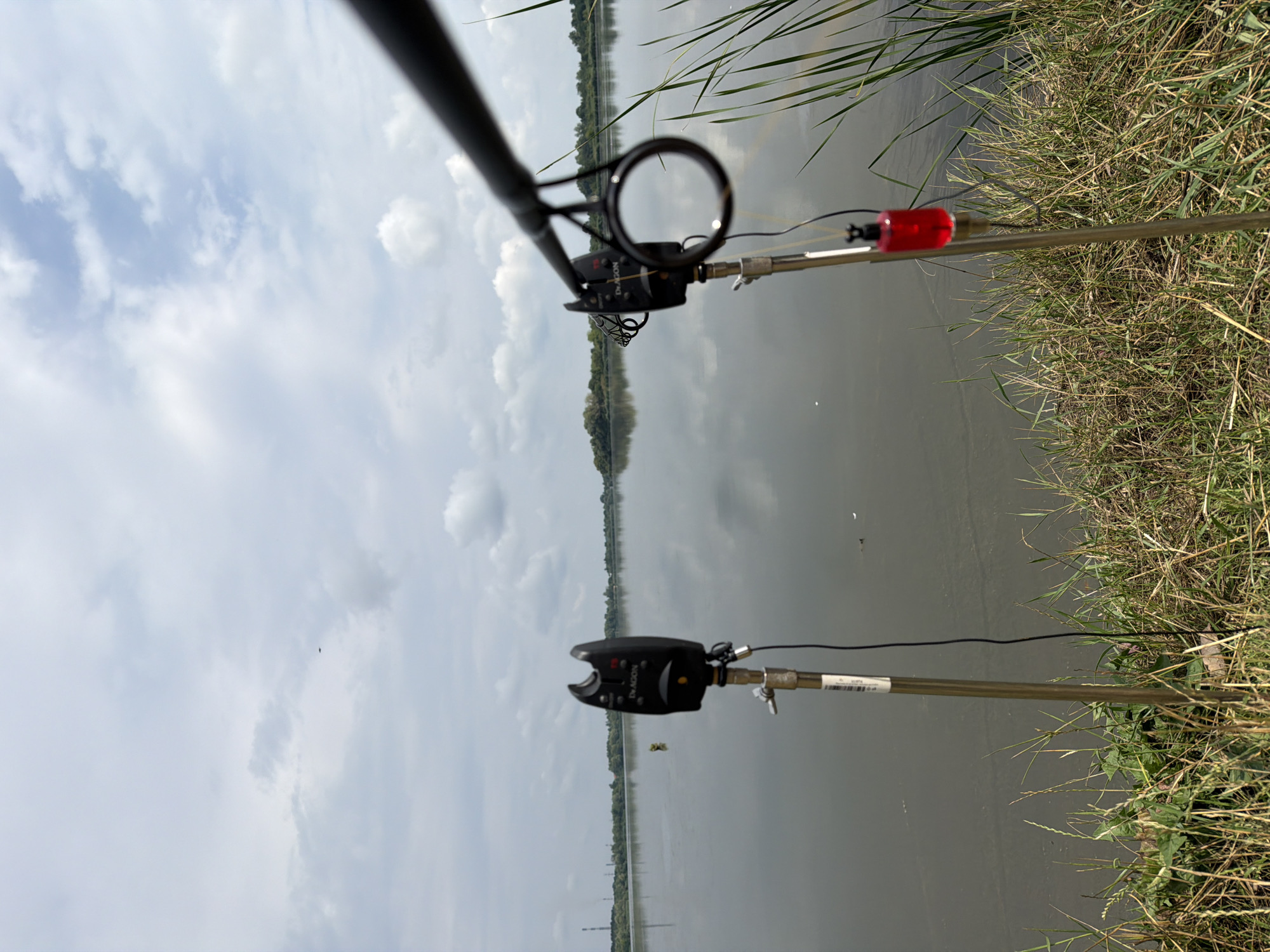 The image shows a calm body of water, likely a lake or river, under a cloudy sky. Two fishing rod pods are set up on the grassy bank, each holding a fishing rod. On the left pod, a black electronic bite alarm is visible, with a fishing line extending from it into the water. On the right pod, another bite alarm is present, along with a red fishing indicator hanging from the rod. A fishing rod extends into the frame from the right side of the image, with its line likely connected to the right pod's setup. The environment suggests a tranquil fishing spot with vegetation along the water's edge. The provided location details state that this image was taken in the city of Ruda, Ukraine.