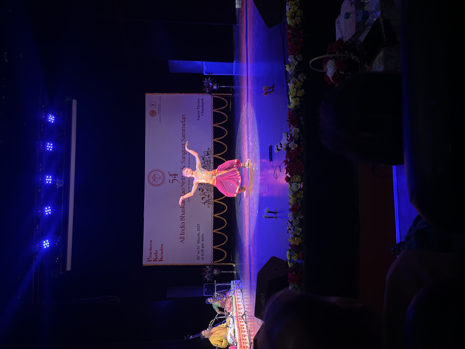 The image shows a stage performance at the Tagore Theatre in Chandigarh, India (latitude: 30.733, longitude: 76.789). A female dancer wearing a pink costume and minimal jewelry is performing on a stage illuminated by spotlights. She has her arms raised in a graceful pose. Behind her is a large banner reading "54th All India Bhaskar Rao Nritya & Sangeet Sammelan". The banner also includes the dates "26th to 31st March 2023" and the time "at 6:30 pm daily". The stage is decorated with a garland of flowers and there are two microphones set up on the stage floor. The image is taken from the audience's perspective, and the silhouette of two spectators can be seen in the foreground. The audience seats are dark and only a few details are visible, including a speaker with the word "JAGJIT" printed on it. The theater's walls are a deep blue color. The ceiling of the auditorium is not visible. The lighting is dim and the stage is lit by bright spotlights.