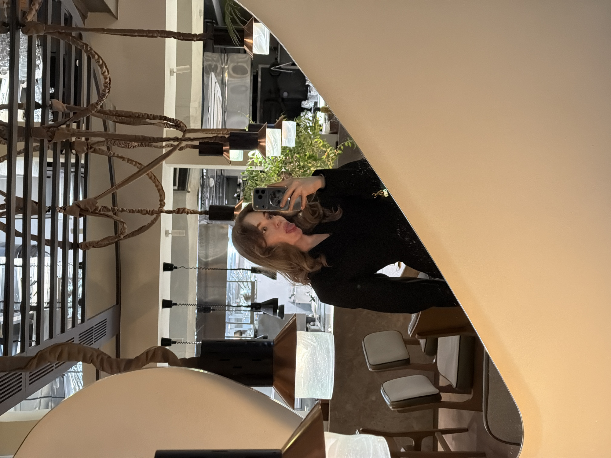 A woman takes a selfie in front of a mirror in what appears to be a restaurant setting. The scene is from Moscow, Russia. The woman is wearing a black long-sleeved top and holds a phone. She is making a playful gesture by sticking out her tongue. Behind her is the restaurant interior, featuring dining tables and chairs, and a unique lighting fixture.