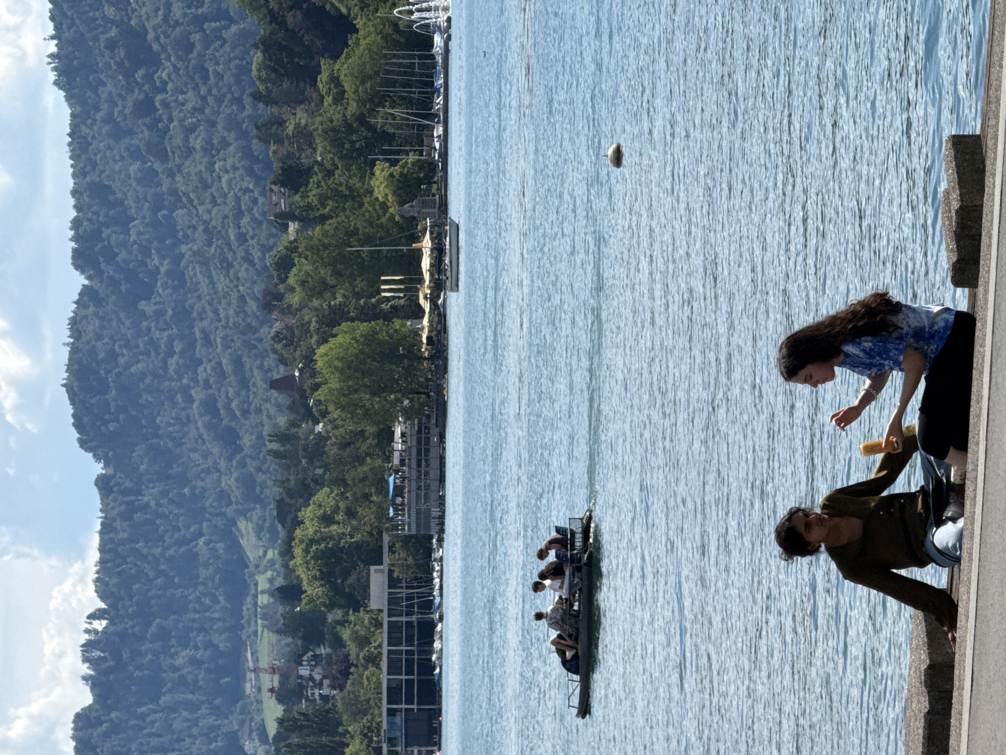 This is a full-frame, eye-level, vertical shot taken on a bright, sunny day at the Zurichsee (Lake Zurich) in Zurich, Switzerland. In the foreground, two young women are sitting on a concrete embankment facing the water. The woman on the left, wearing a dark green long-sleeved shirt and light blue pants, is looking towards the woman on the right. The woman on the right, wearing a blue and white tie-dye t-shirt and black pants, is holding a yellow cylindrical object, possibly a snack or drink, and appears to be interacting with the woman on the left.
In the middle ground, a small boat with four people is being pedaled across the blue water. The people in the boat are seated and appear to be enjoying a leisurely ride.
The background features a cityscape with modern buildings, including a long, rectangular structure with windows and a pier with boats docked. Further in the distance, a lush, tree-covered hill rises, indicating the natural landscape surrounding the lake. The sky is blue with scattered white clouds. A white buoy is visible in the water between the foreground and the boat.