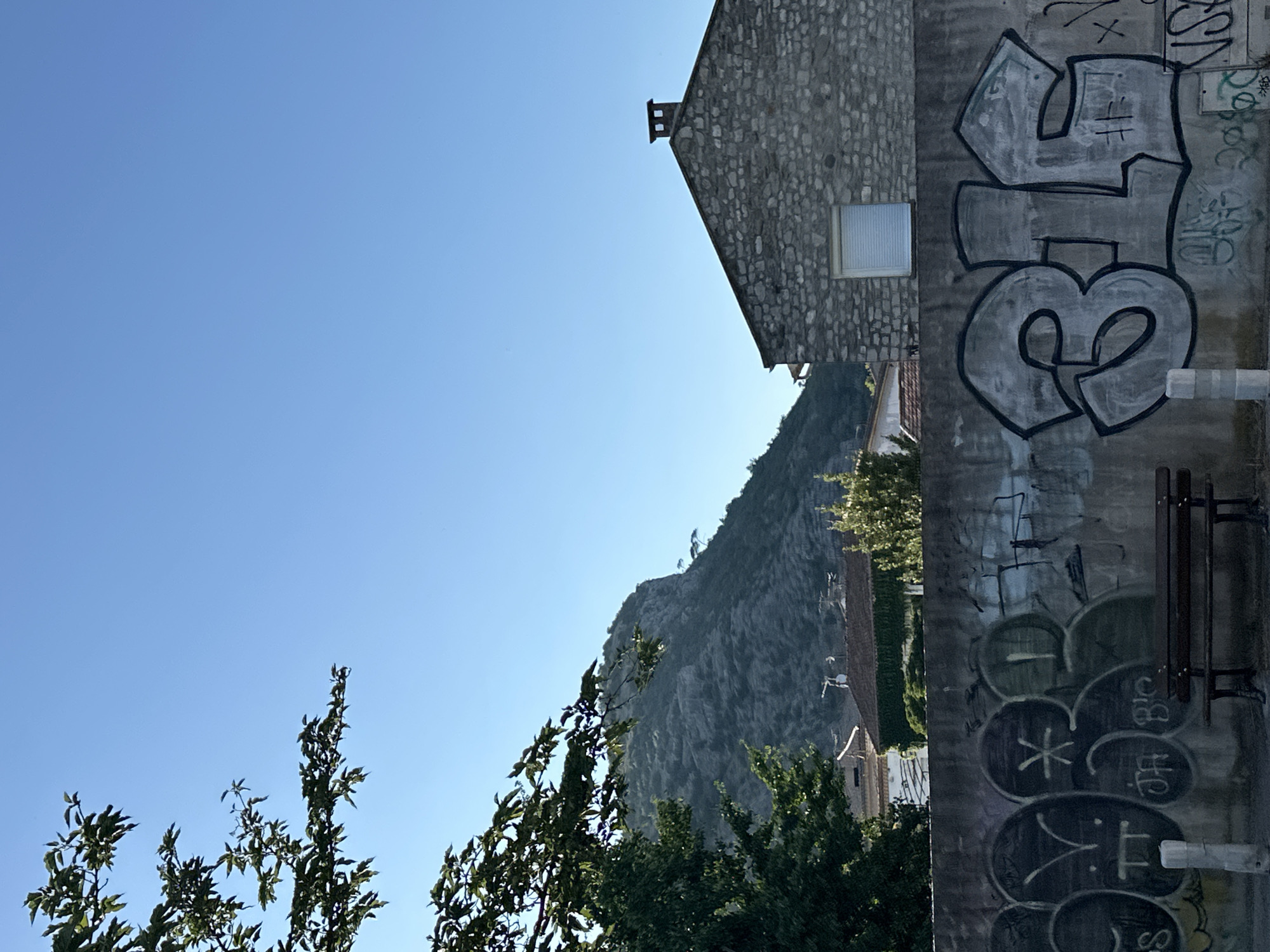 The media file is a vertical, outdoor photograph taken on a clear, sunny day. The image is dominated by a bright blue sky on the left, which transitions to a steep, rocky mountainside on the right. In the foreground, the edge of a building with a stone facade is visible, featuring a small window and a chimney. A section of a concrete wall on the right side of the image is covered in graffiti, with various tags and bubble letters in grey and black. A dark wooden bench is mounted on this wall. Below the mountainside, glimpses of houses and lush green trees can be seen. The overall impression is one of a bright, clear day in a mountainous area with an urban element featuring graffiti. The time of day appears to be late morning or early afternoon due to the bright sunlight and clear sky.