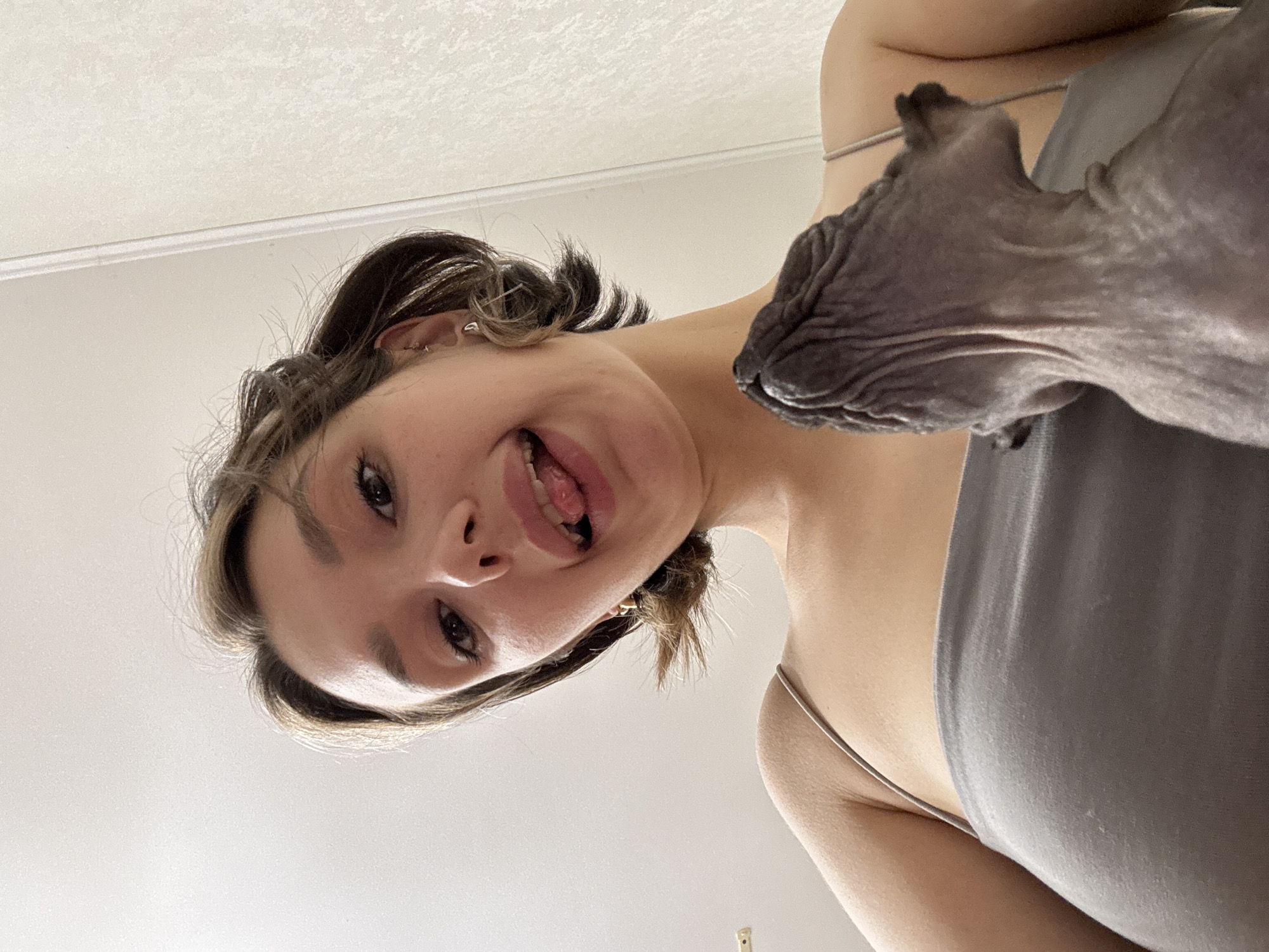 A low-angle selfie shows a young woman with short brown hair and a grey tank top, sticking her tongue out playfully at the camera. A hairless cat is visible in the foreground, with its head turned away from the camera, looking up at the woman. The background consists of a plain wall and ceiling. The scene appears to be indoors, in what looks like a home environment.