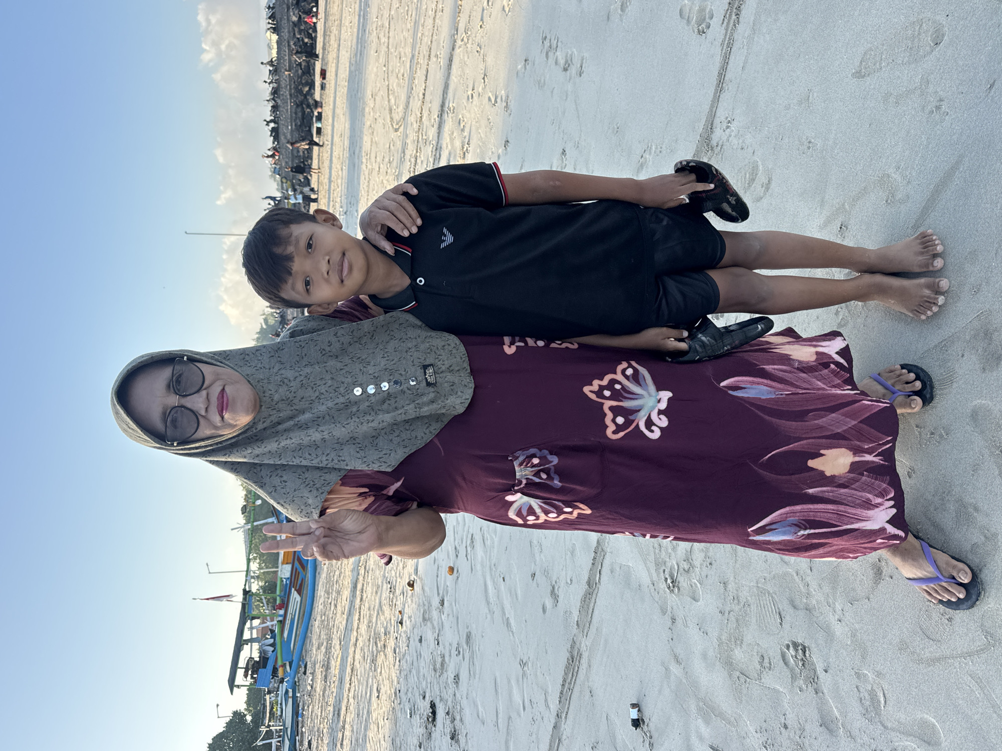 An elderly woman and a young boy stand on a beach in Kuta, Indonesia. The woman, wearing a green hijab, glasses, and a dark red floral dress, is making a peace sign with her left hand. The boy, in a black polo shirt and shorts, has his arm around the woman's shoulders. They are both barefoot, and the boy is holding a pair of black shoes. In the background, there are several colorful fishing boats on the sand and in the shallow water, with some people and motorbikes visible further down the beach. The sky is clear and blue with some clouds.