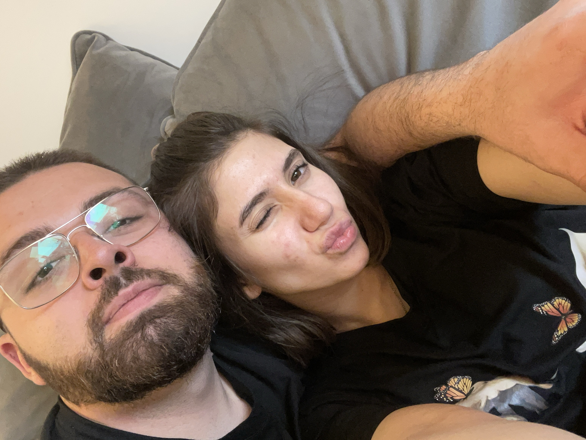 The image shows a selfie of a couple lying down, likely on a couch, given the grey cushion visible in the background. The man on the left has a beard and glasses. The woman on the right is winking and puckering her lips. She's wearing a black t-shirt with butterfly prints. There is not enough information to determine if the image was taken in Sofia, Bulgaria.