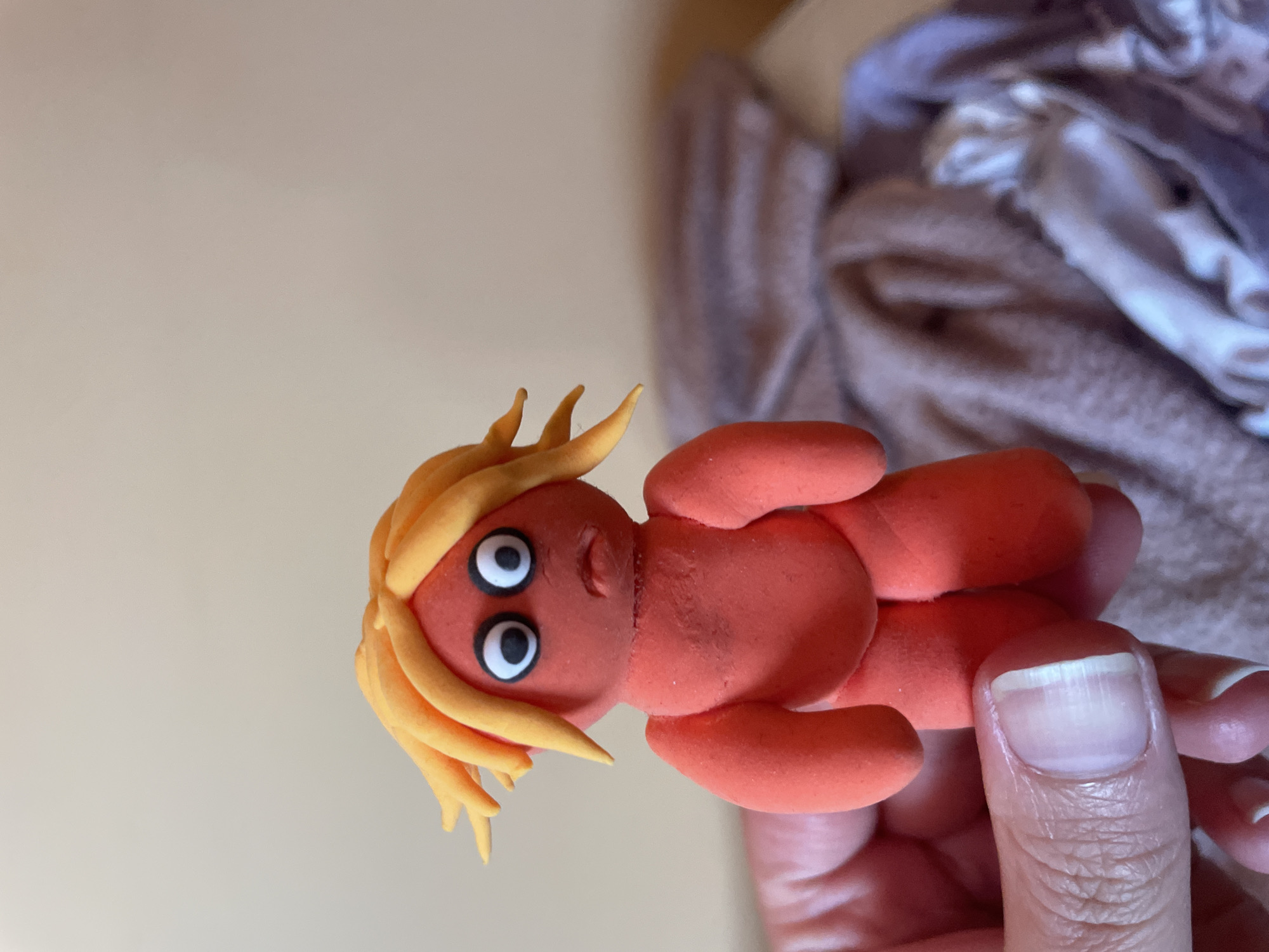 A clay figurine is being held in a person's hand. The figurine has an orange body, yellow hair, and large white eyes with black pupils. The hand holding the figurine has a visible thumbnail with a healthy appearance. The background consists of a light beige wall and a blurry area of what appears to be draped fabric or clothing. The location is unknown.