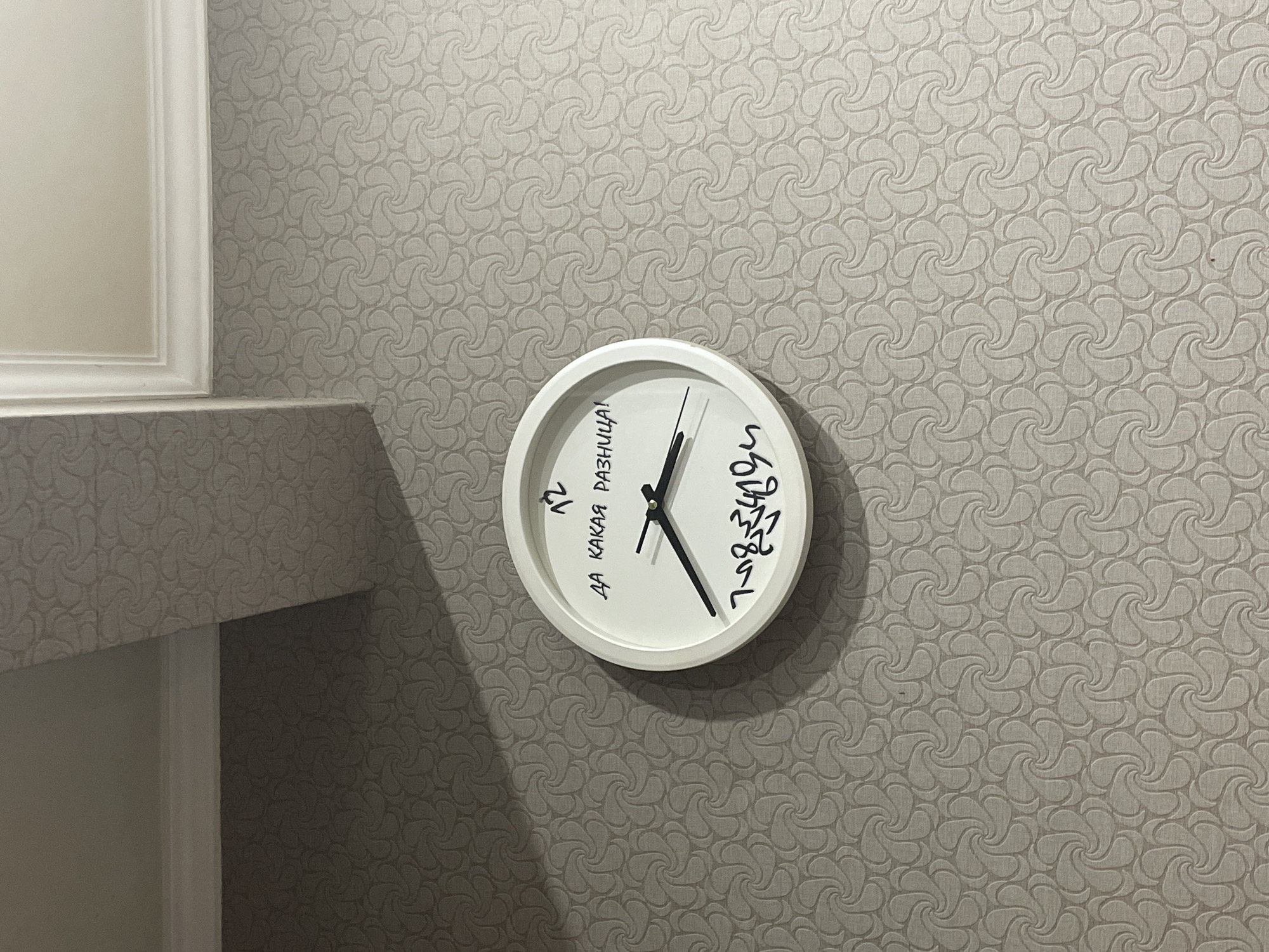 A white analog clock with black hands hangs on a textured gray wall. The clock face has the number "12" at the top, and the phrase "ДА КАКАЯ РАЗНИЦА!" is written in black cursive text below it. Below that, there is a drawing or signature in black cursive. The clock is casting a shadow to the left. The wall has a repeating swirling pattern. The image appears to be taken indoors. There is no indication of the city of Kaliningrad, Russia, or any specific location.