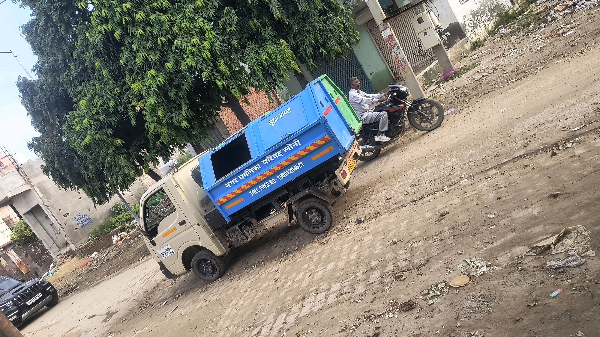 In Loni, India, on a dirt road, a beige truck with a bright blue garbage bin is parked. The truck has a small cab and a large rear bin that is mostly blue with some green at the top and yellow and orange stripes on the side. Text on the bin reads "सूखा कचरा" (Dry Waste) and "नगर पालिका परिषद, लोनी" (Nagar Palika Parishad, Loni), along with a toll-free number "TOLL FREE NO- 18001204621". A man wearing a light-colored shirt and pants is riding a black and red motorcycle with a black helmet, moving past the truck. To the left, a black SUV with a license plate that reads "UP14 G07474" is parked. The environment appears to be a somewhat unkempt urban area with some debris on the ground and buildings in the background. A large tree with dense green foliage is visible behind the truck.