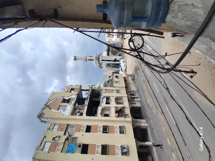 An overhead photograph captured from a building balcony depicts a devastated urban street scene in Khan Yunis, Palestinian Territory, under an overcast sky. In the immediate foreground on the right, a large blue plastic water dispenser bottle sits on a concrete ledge, with numerous dark, loose electrical wires extending across the frame, attached to a vertical support pole.
The midground features a two-lane asphalt road with faded white markings. A person in dark attire with a light head covering walks on the left sidewalk, moving away from the viewer. On the right side of the road, near a debris-strewn area, another small figure is discernible.
The left side of the frame is dominated by a severely damaged multi-story building with a light beige facade. Significant sections of its upper and middle floors are destroyed, exposing internal structures and rubble. Some windows are missing, while others are covered with blue tarps. Further in the background, a large mosque with a damaged dome and a broken minaret stands partially collapsed, surrounded by debris. Additional damaged structures are visible beyond the mosque. The overall environment suggests extensive structural destruction.